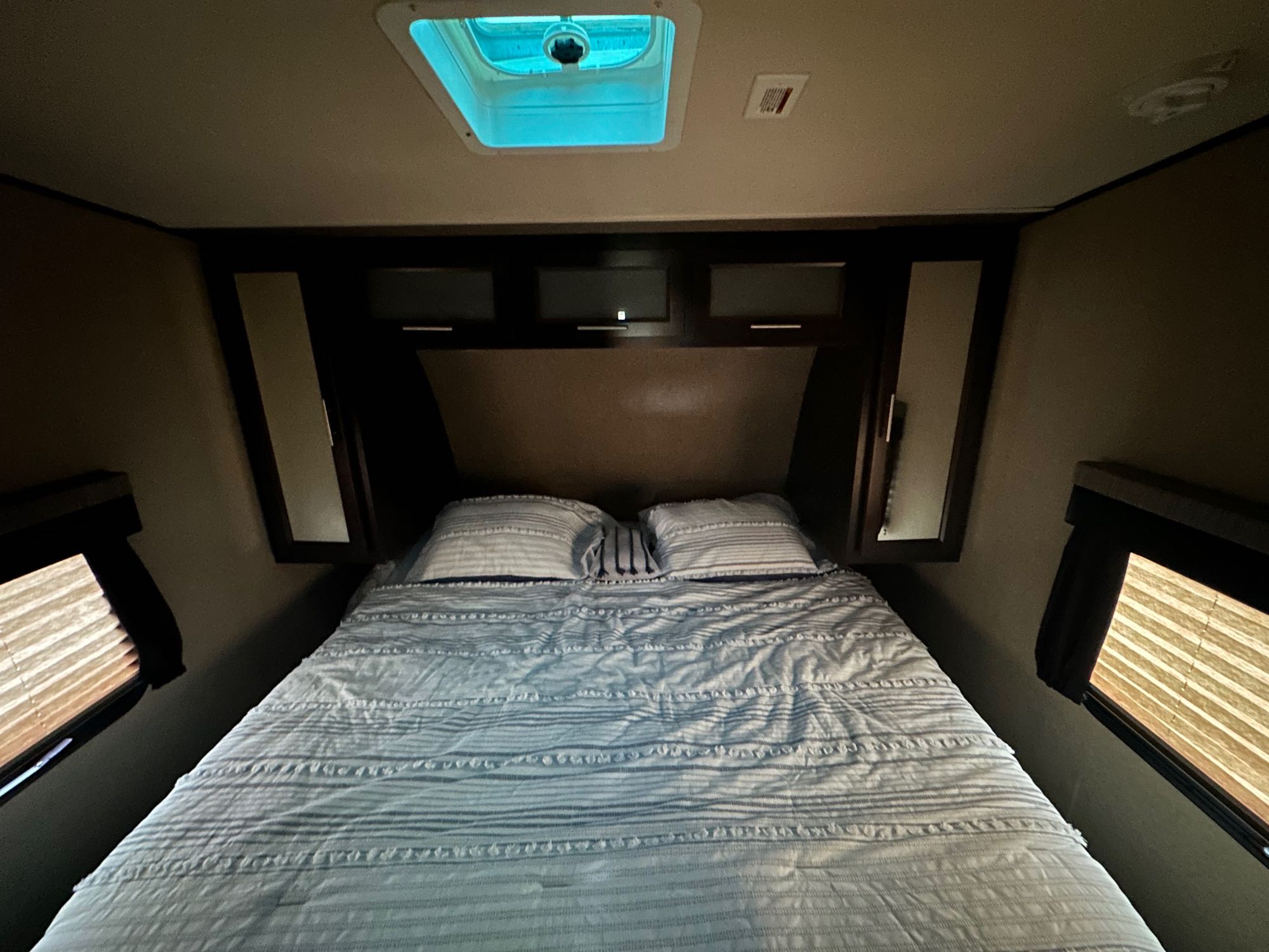 Cozy RV rental bedroom: striped bed, overhead cabinets, and side windows with blinds. Blue ceiling vent adds ambiance for travel.. Forest River Nitro Xlr 2018