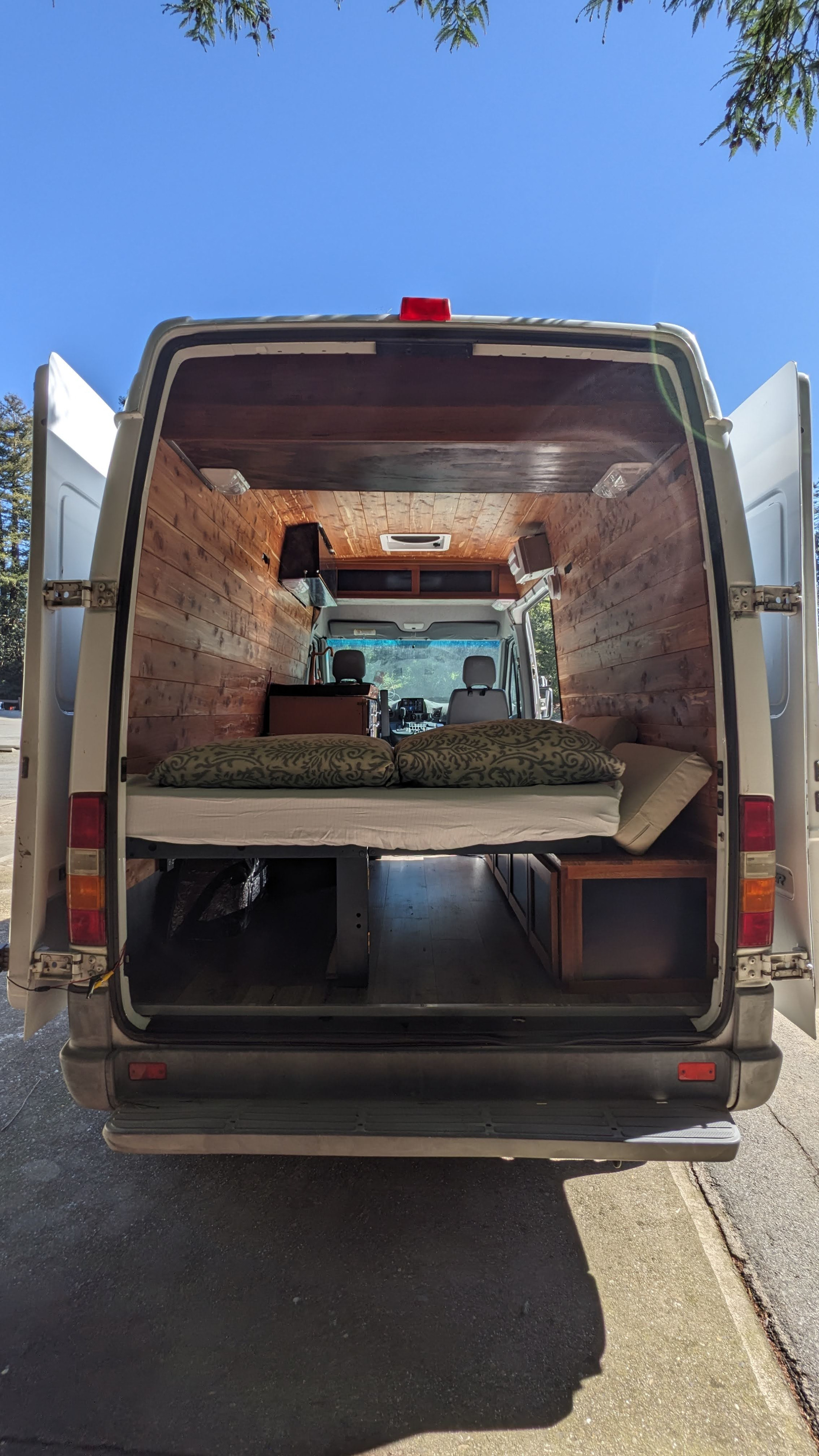 This RV rental showcases a bright, wood-paneled camper van interior with a comfortable bed, under-bed storage, and front cab view.. Dodge Sprinter Van 2005