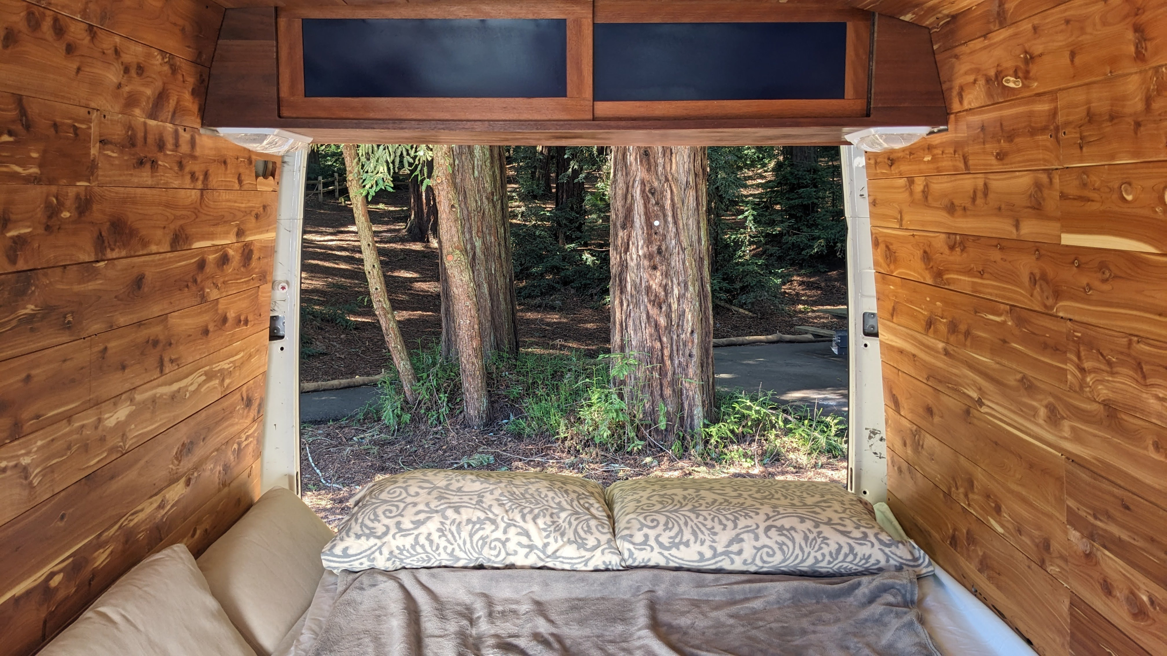 Cozy rv rental camper interior, looking out the open back to a serene forest with large trees, wood-paneled walls, and a comfy bed.. Dodge Sprinter Van 2005