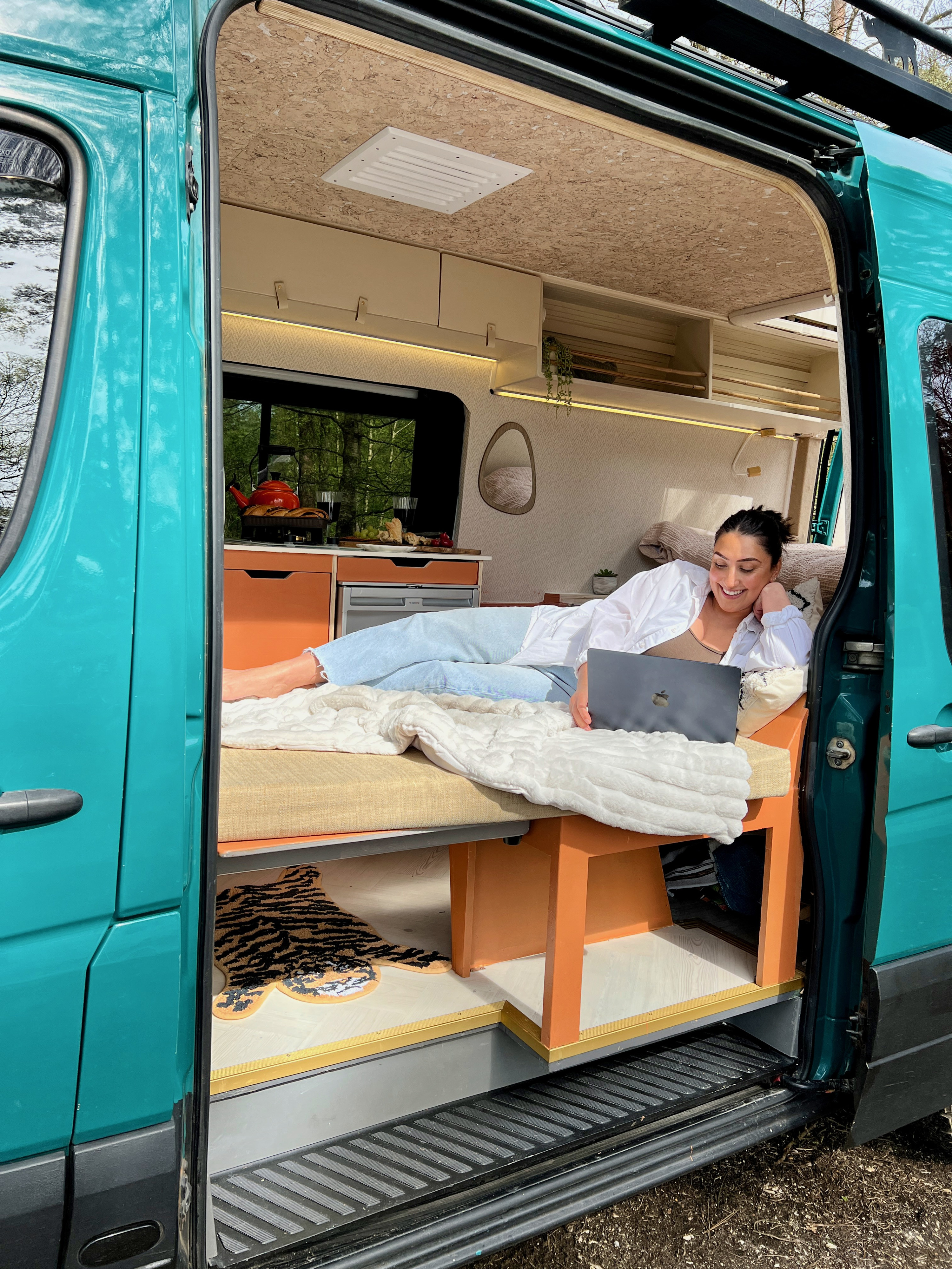 A woman works on her laptop inside a teal **rv rental**, lounging on a cozy bed with a full kitchen visible.. Mercedes-Benz Sprinter 2016