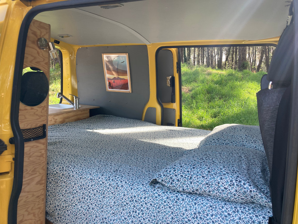 Inside a yellow RV rental: a comfortable bed, small sink, and open back doors showing a sunny, grassy field.. Volkswagen Transporter T5 2012