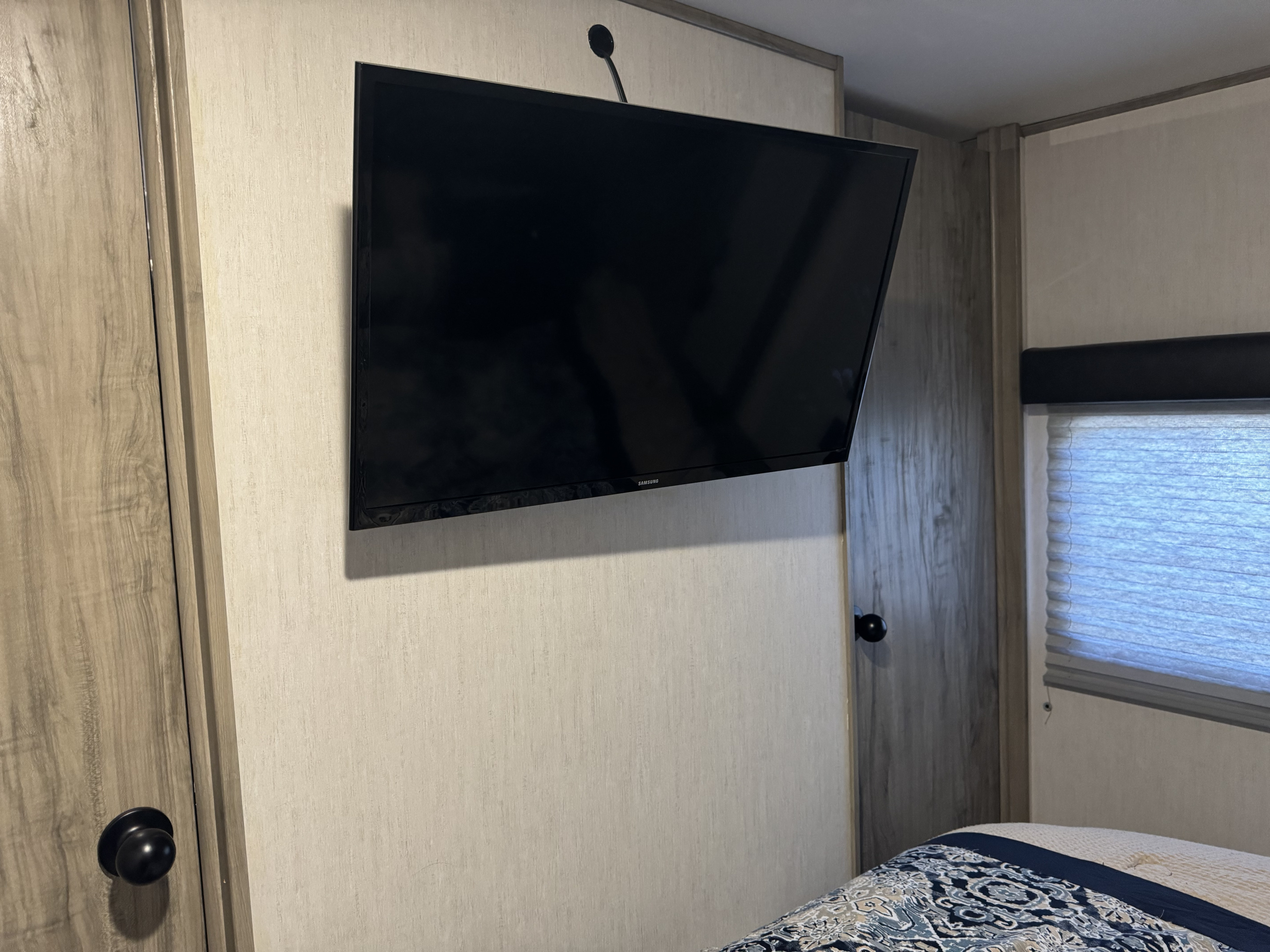 Compact RV rental bedroom showing a wall-mounted TV, two light wood doors, and a window with blinds above a bed.. Forest River Cherokee Alpha Wolf 2022