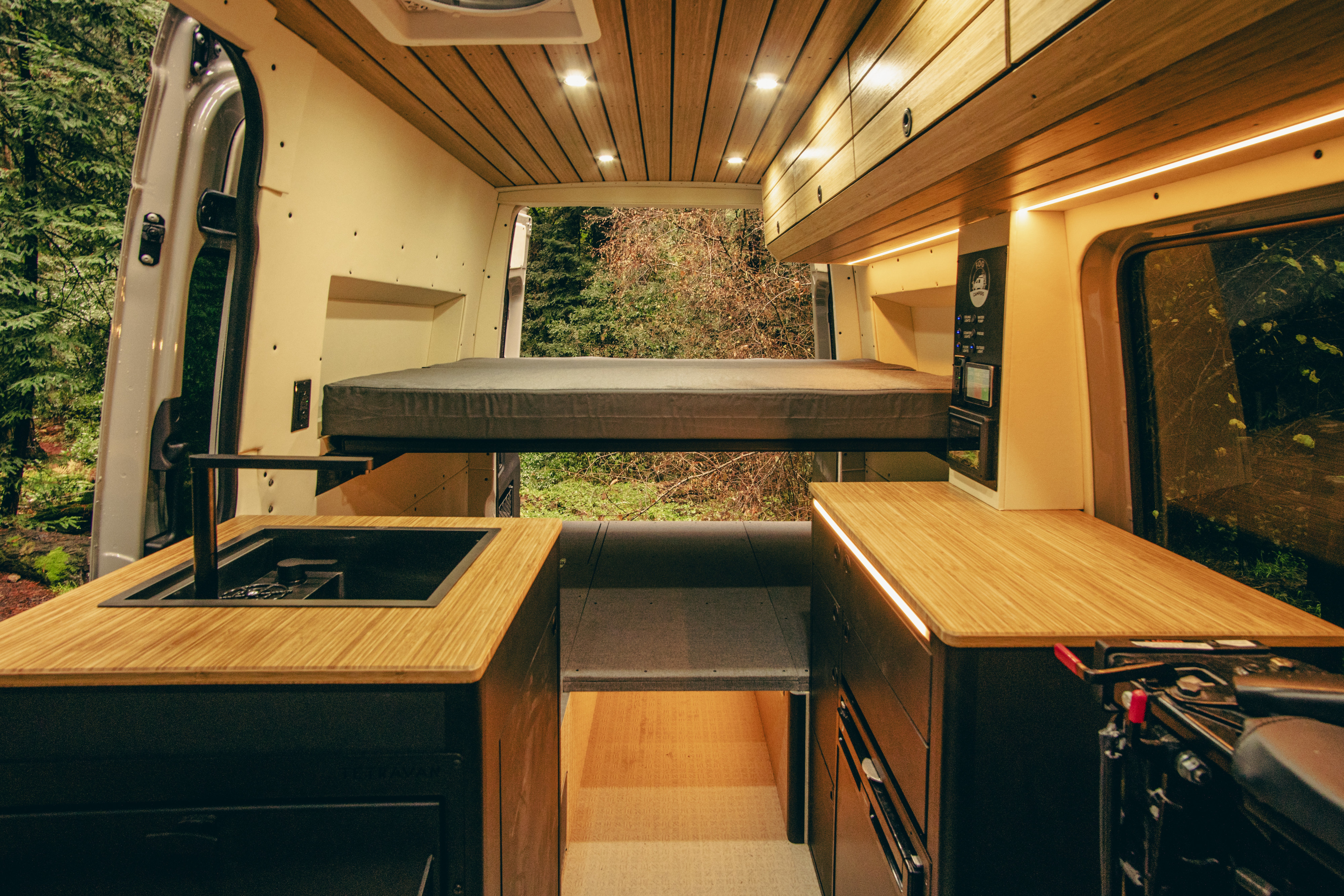 Inside a custom RV rental, this spacious camper van features a raised bed, kitchen, and forest views from the open rear.. Mercedes-Benz Sprinter 2023
