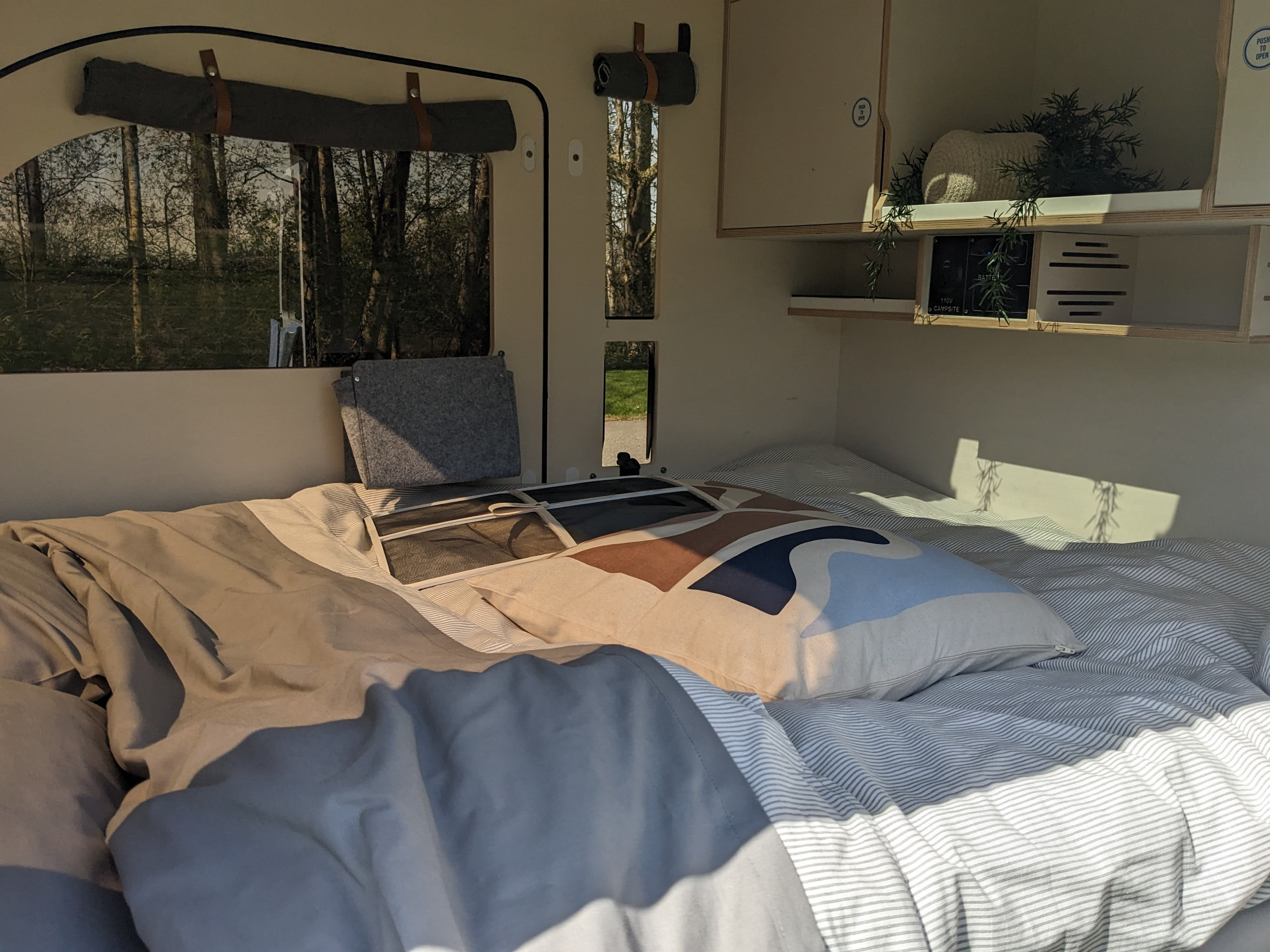 RV rental bedroom interior: bed with striped bedding and decorative pillow, windows with tree views, and shelving.. droplet XL 2023