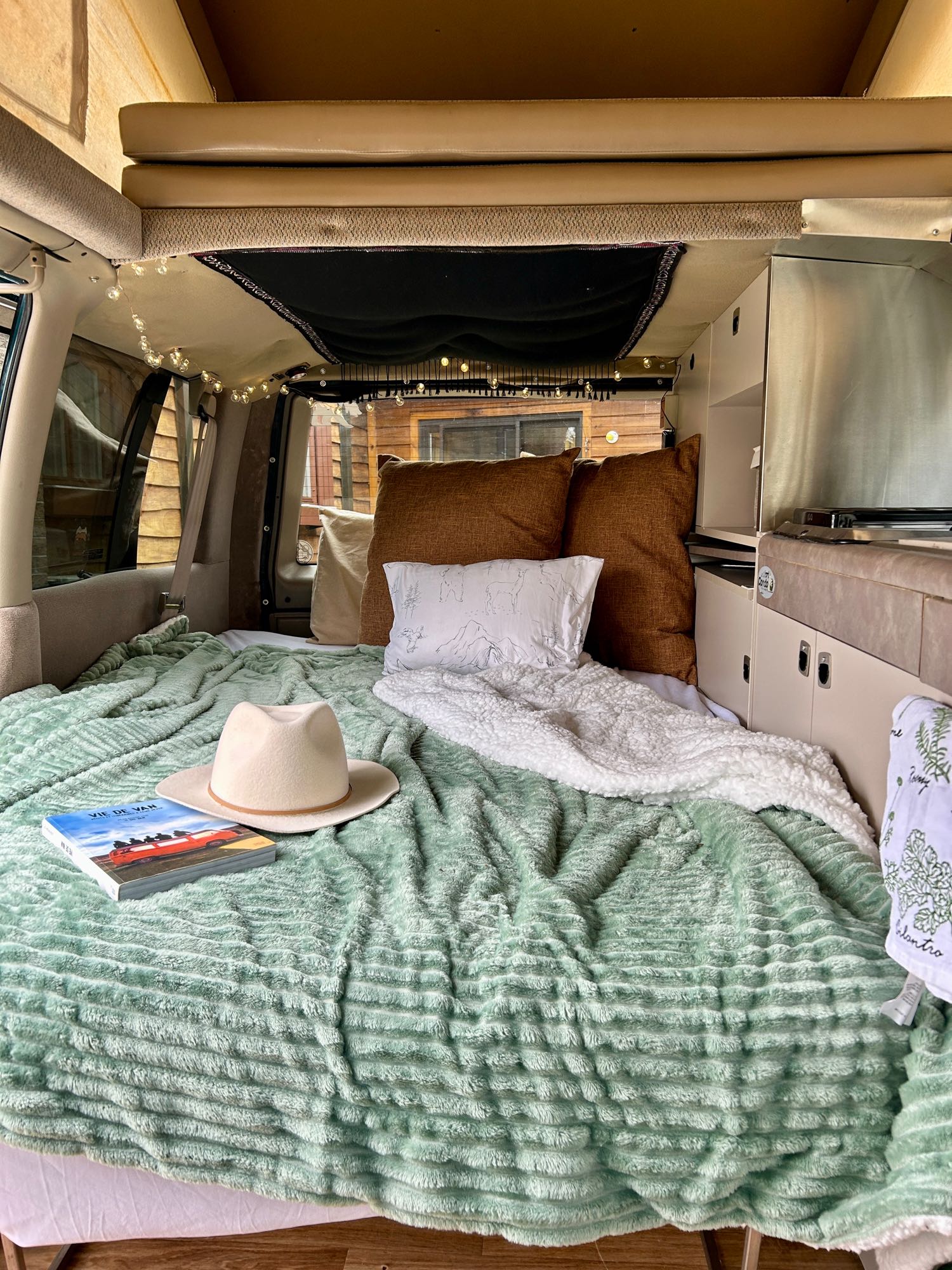 Inside a cozy RV rental: a comfortable bed with green and white blankets, a book, hat, and string lights, ready for travel.. Chevrolet Astro 1997