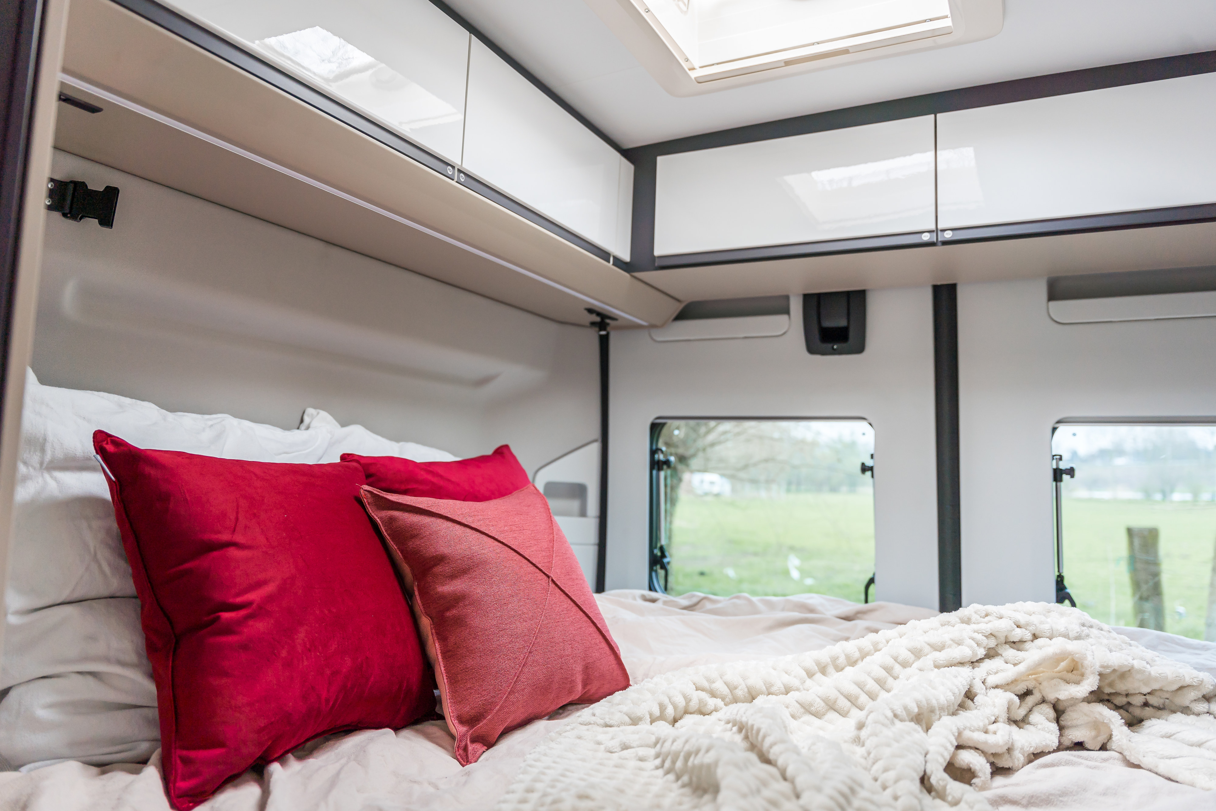Cozy RV rental bedroom: soft white bedding, red pillows, overhead storage, and windows overlooking green fields.. Adria TWIN PLUS SPB 2024