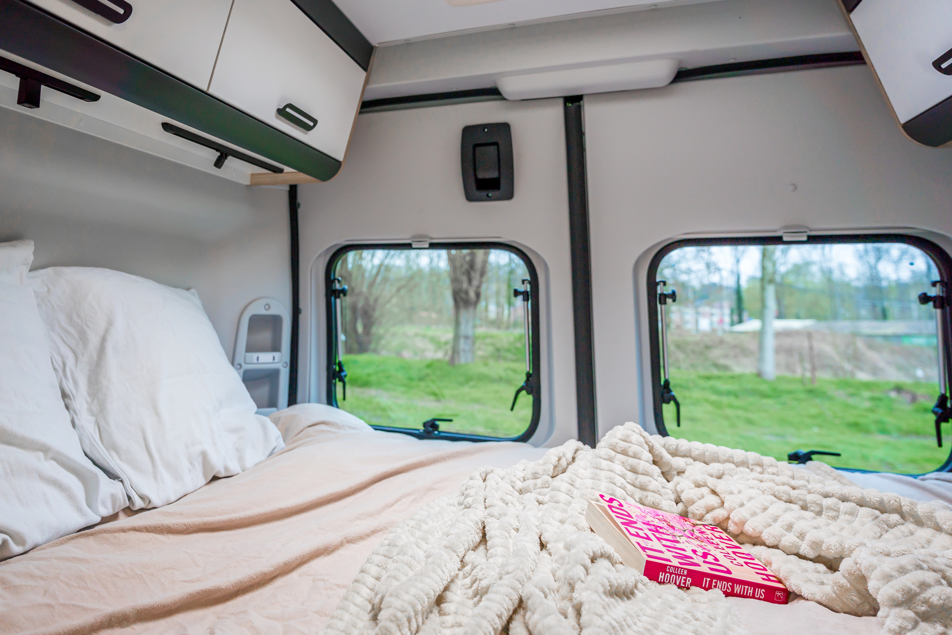 Inside a comfy rv rental: a cozy bed with pillows, blanket, and a book "It Ends With Us," overlooking a green view.. SUN LIVING SP 60 AVEC TENTE DE TOÎT 2024
