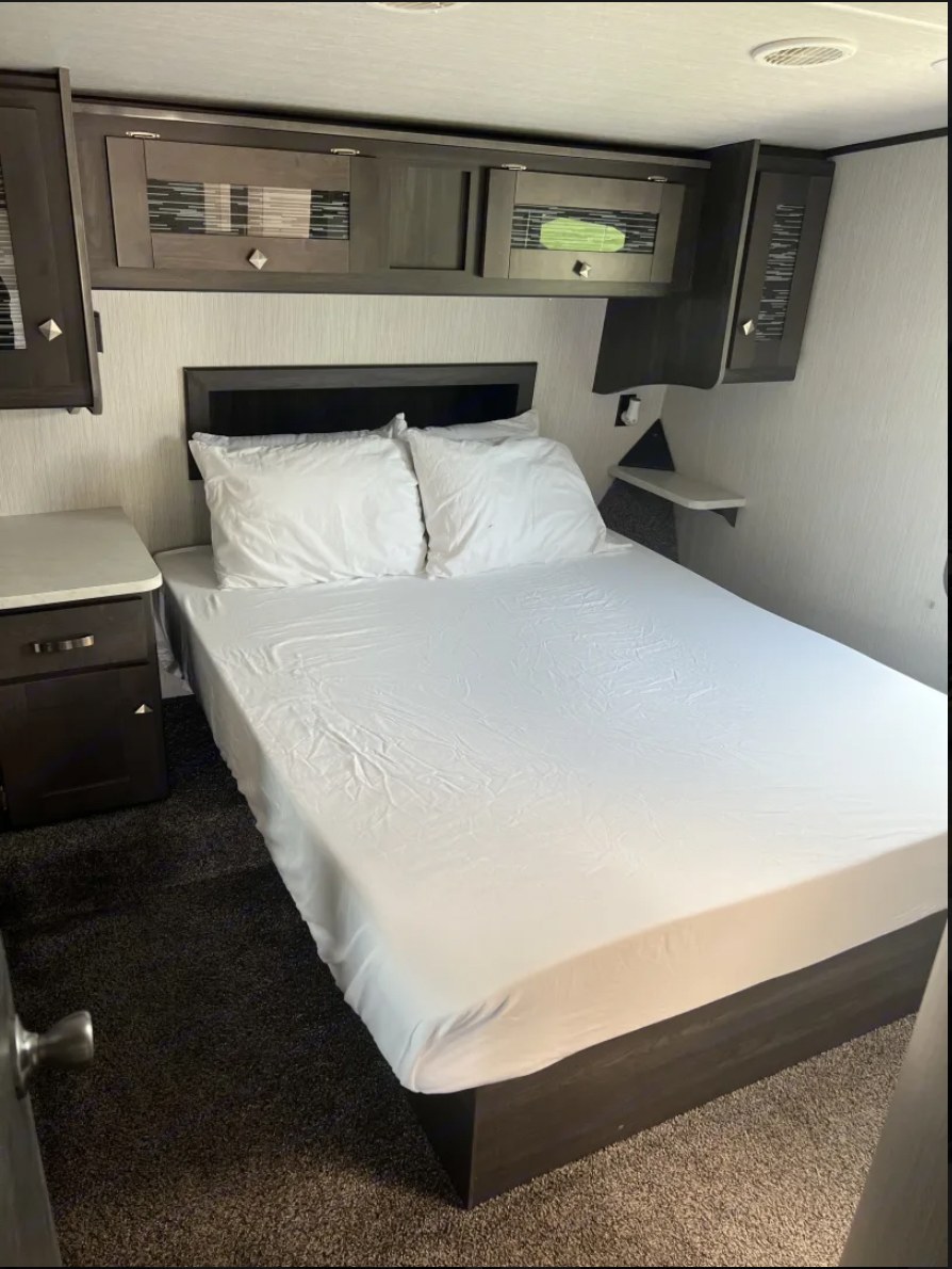 This comfortable RV rental bedroom features a full bed with white linens, overhead storage cabinets, and a compact nightstand.. Heartland RVs Fuel Toy Hauler 2021