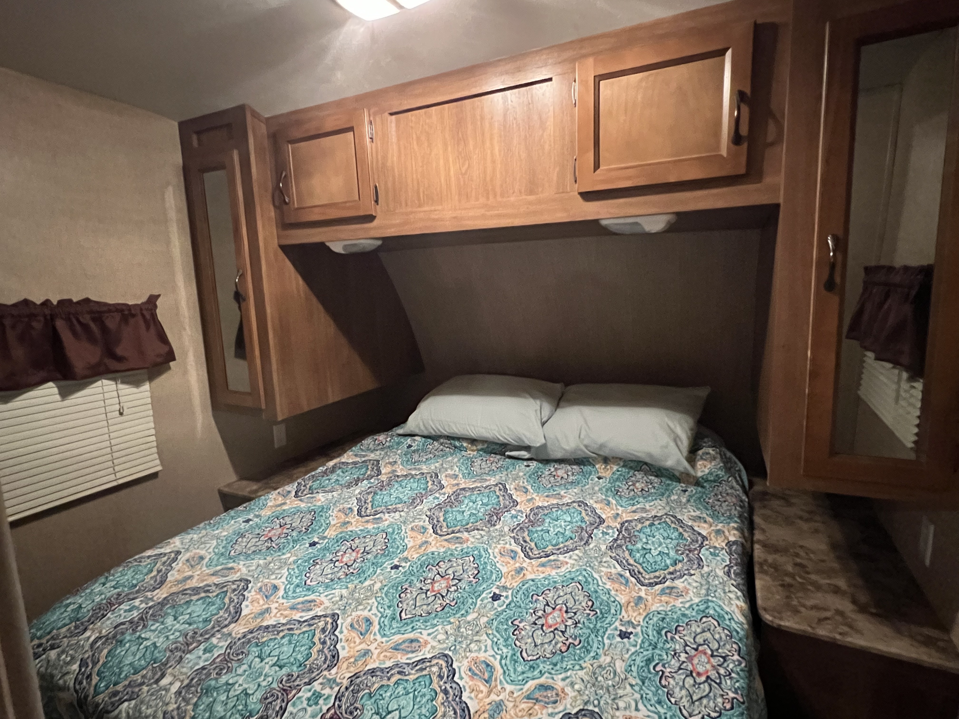 Inside a comfy RV rental bedroom: bed with patterned comforter, wooden cabinets, and window. Ideal for travel.. Coachmen Catalina 293QBCK 2015