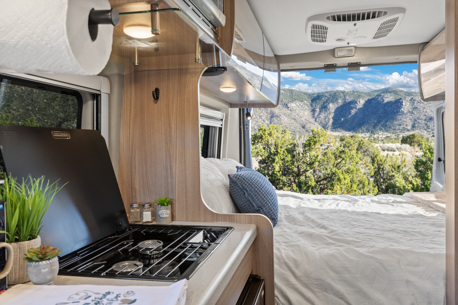 Inside an RV rental: a compact kitchen, bed, and open rear doors revealing a scenic mountain landscape.. Thor Sequence 2022