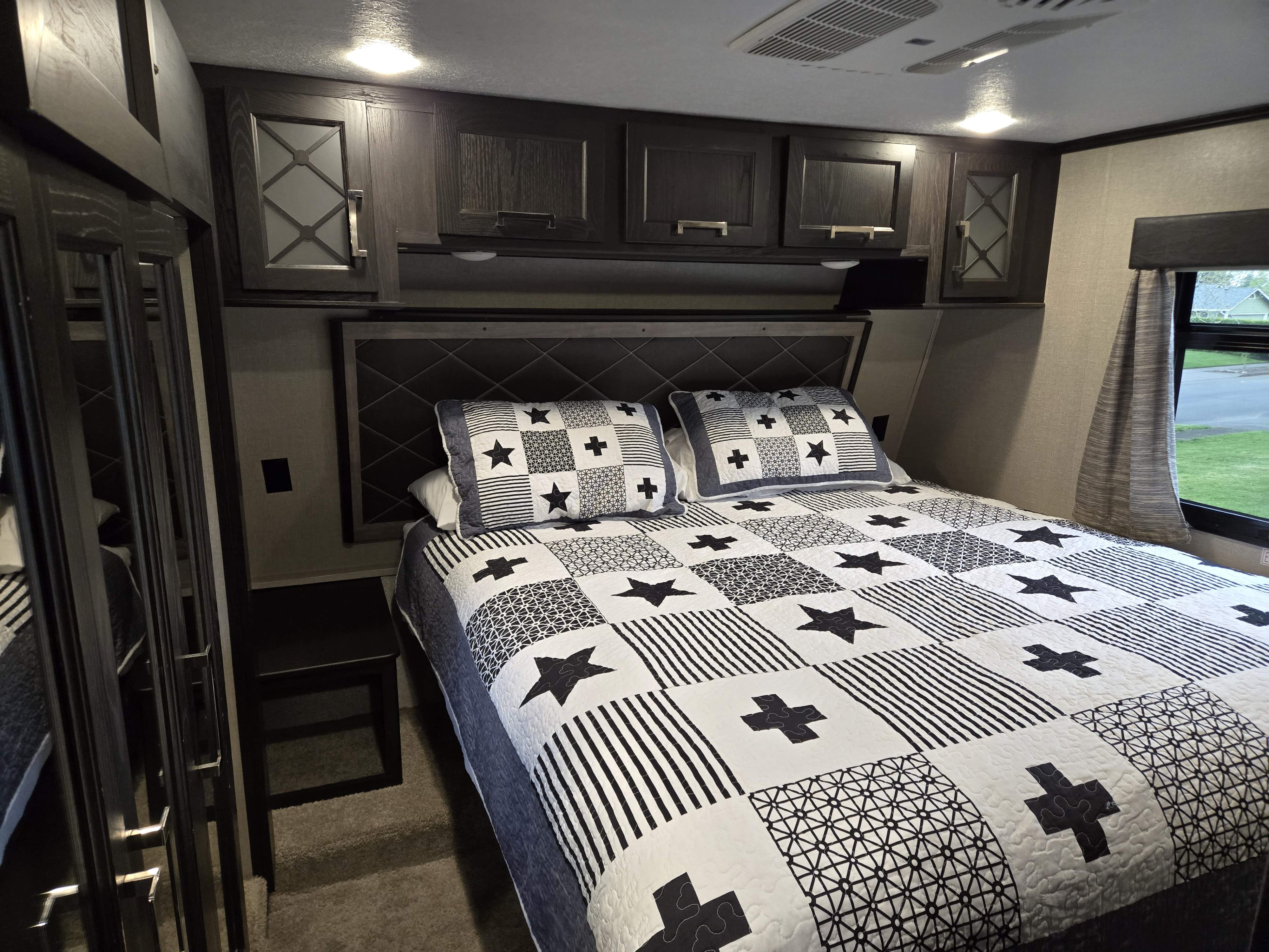 Relax in this RV rental bedroom: a black & white patterned quilt on the bed, overhead cabinets, and a window to the right.. Keystone Fuzion 2018