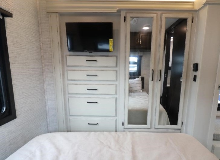 RV rental bedroom: a mounted TV above five white drawers, plus mirrored closet doors reflecting the RV interior. Cozy bed in foreground.. Entegra Vision 2024