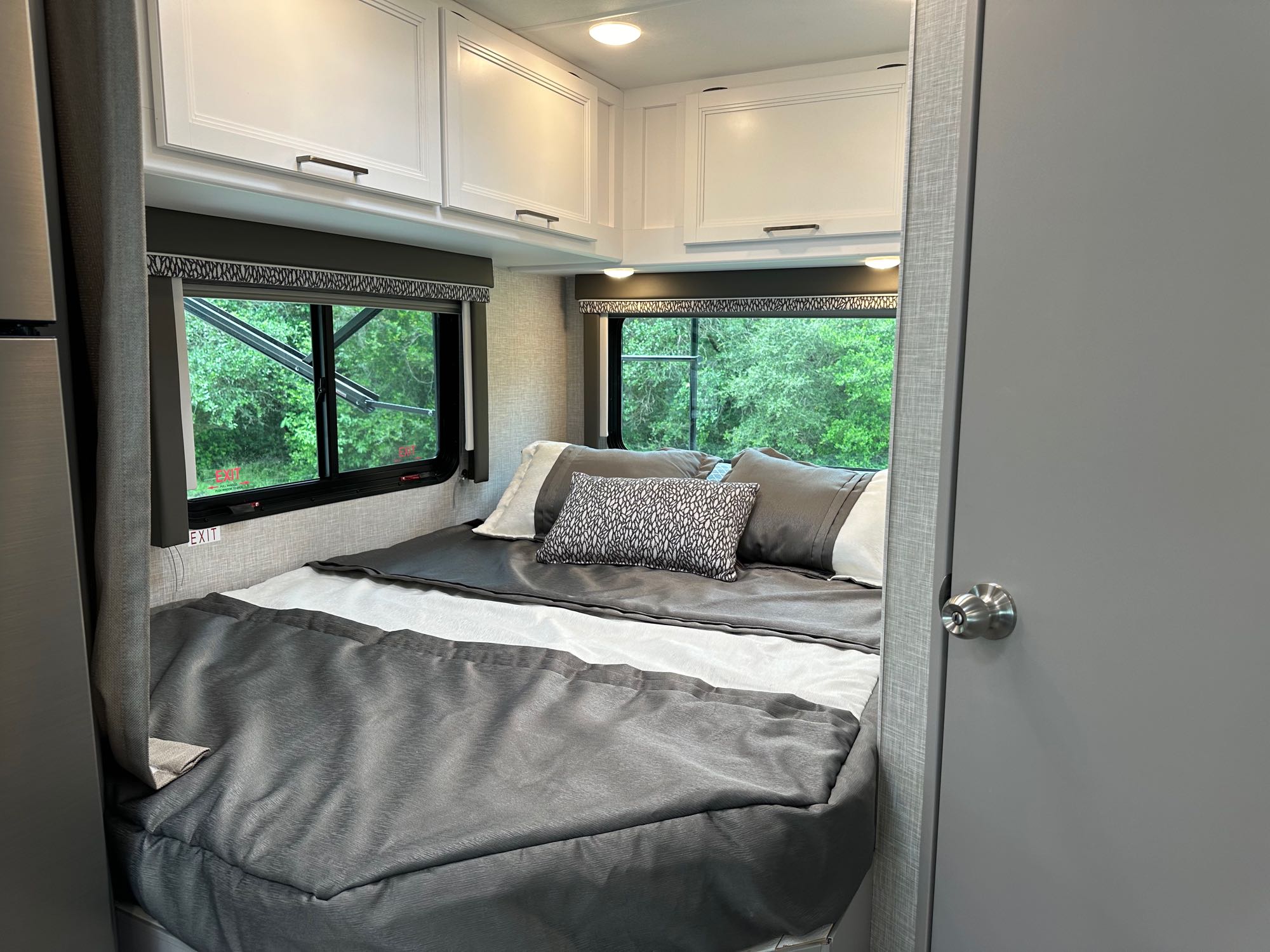 Interior of an RV rental bedroom showing a bed with grey bedding, white overhead cabinets, and lush green trees visible through windows.. Thor Quantum LC 2024