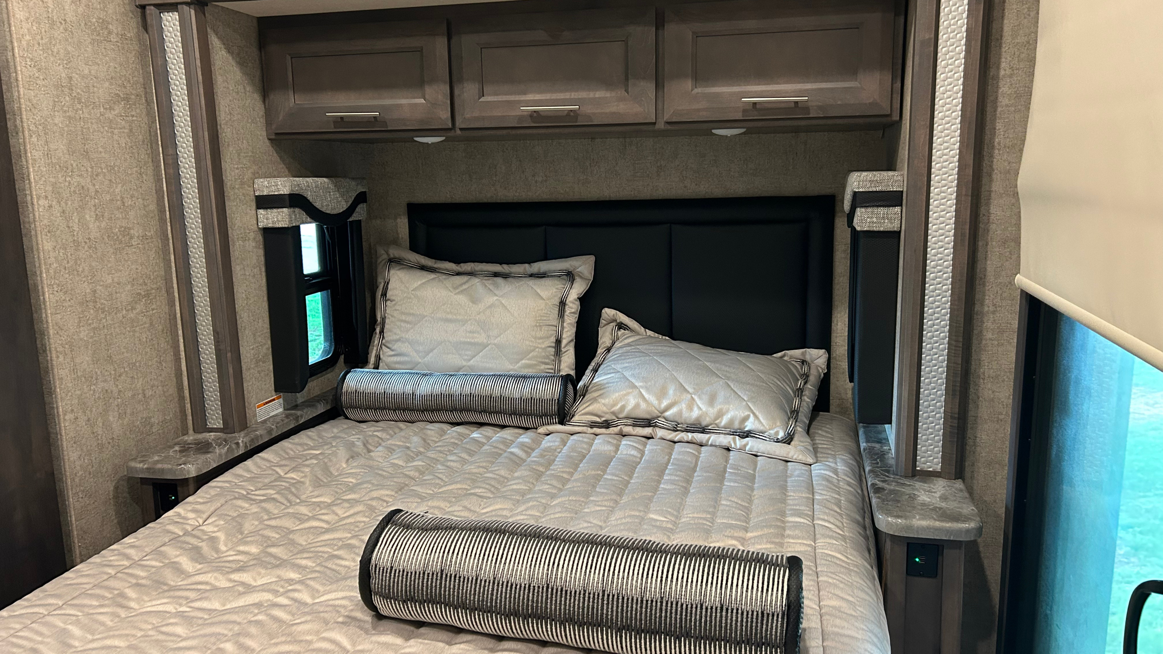 An RV rental bedroom showing a bed with grey bedding, black headboard, overhead cabinets, and side shelves with USB ports.. Thor Outlaw 2024