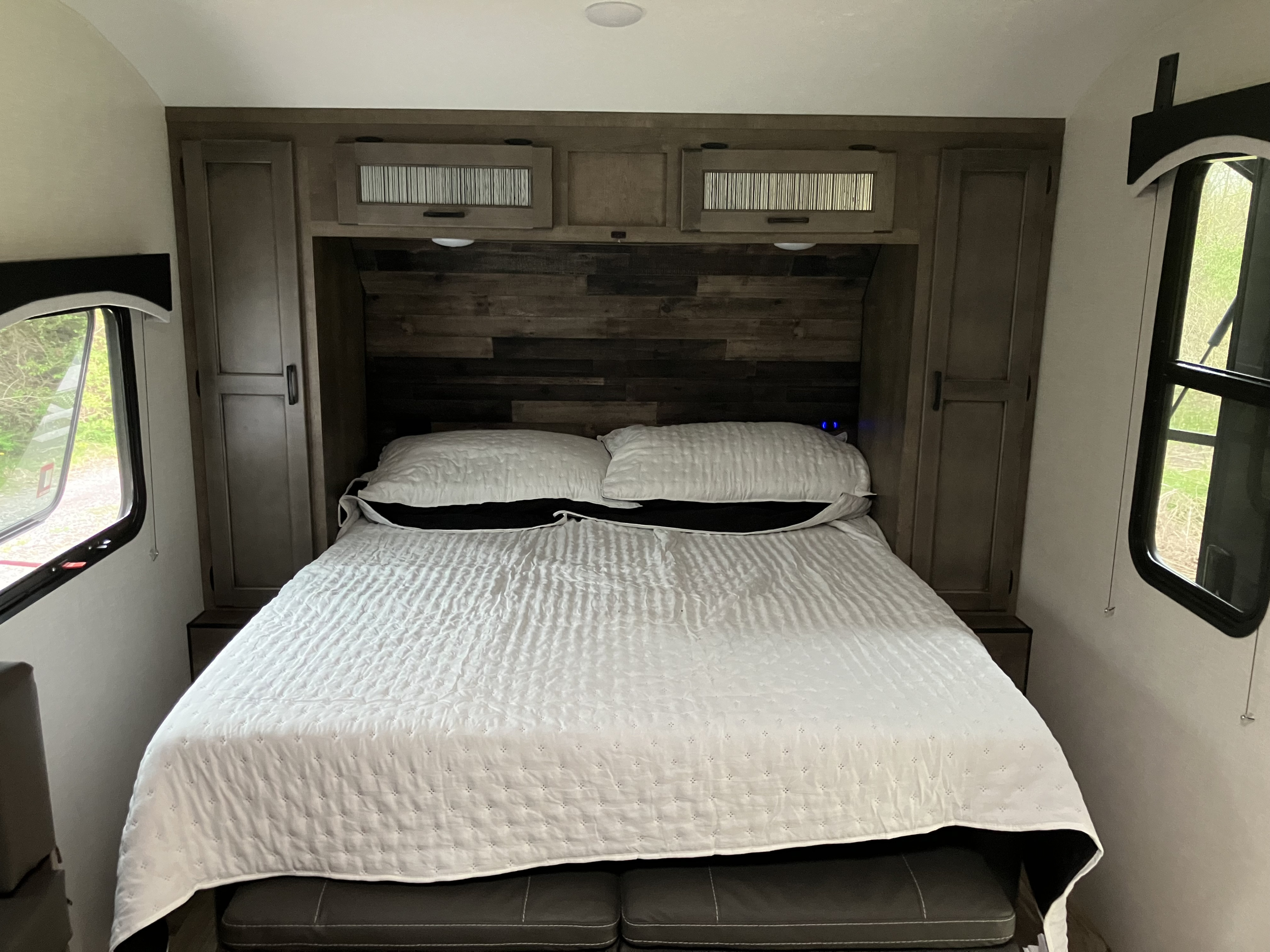 Cozy RV rental bedroom features a comfy bed, wood headboard, overhead cabinets, and bright windows. Your travel retreat.. Sonic Sonic Trailer 2023