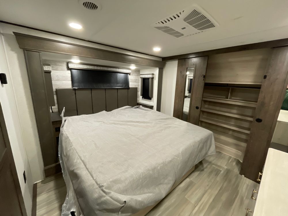 This modern RV rental bedroom features a large bed with a headboard, built-in storage wardrobe, and overhead AC unit for comfortable travel.. Alliance Paradigm 2024