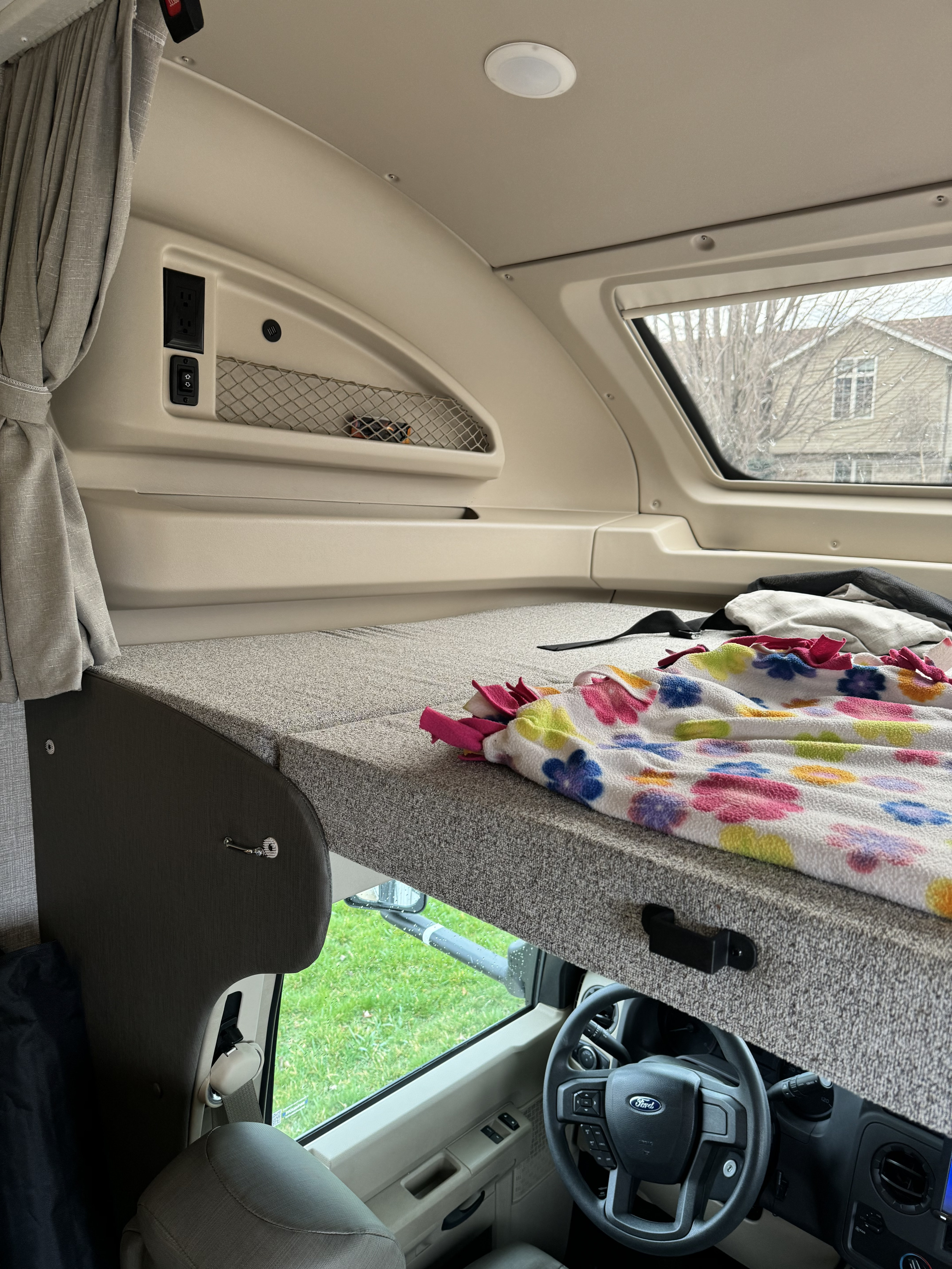 Cozy cab-over bunk in an RV rental, featuring a bed with a colorful blanket, outlets, and the Ford steering wheel below.. Entegra Coach Odyssey 2022
