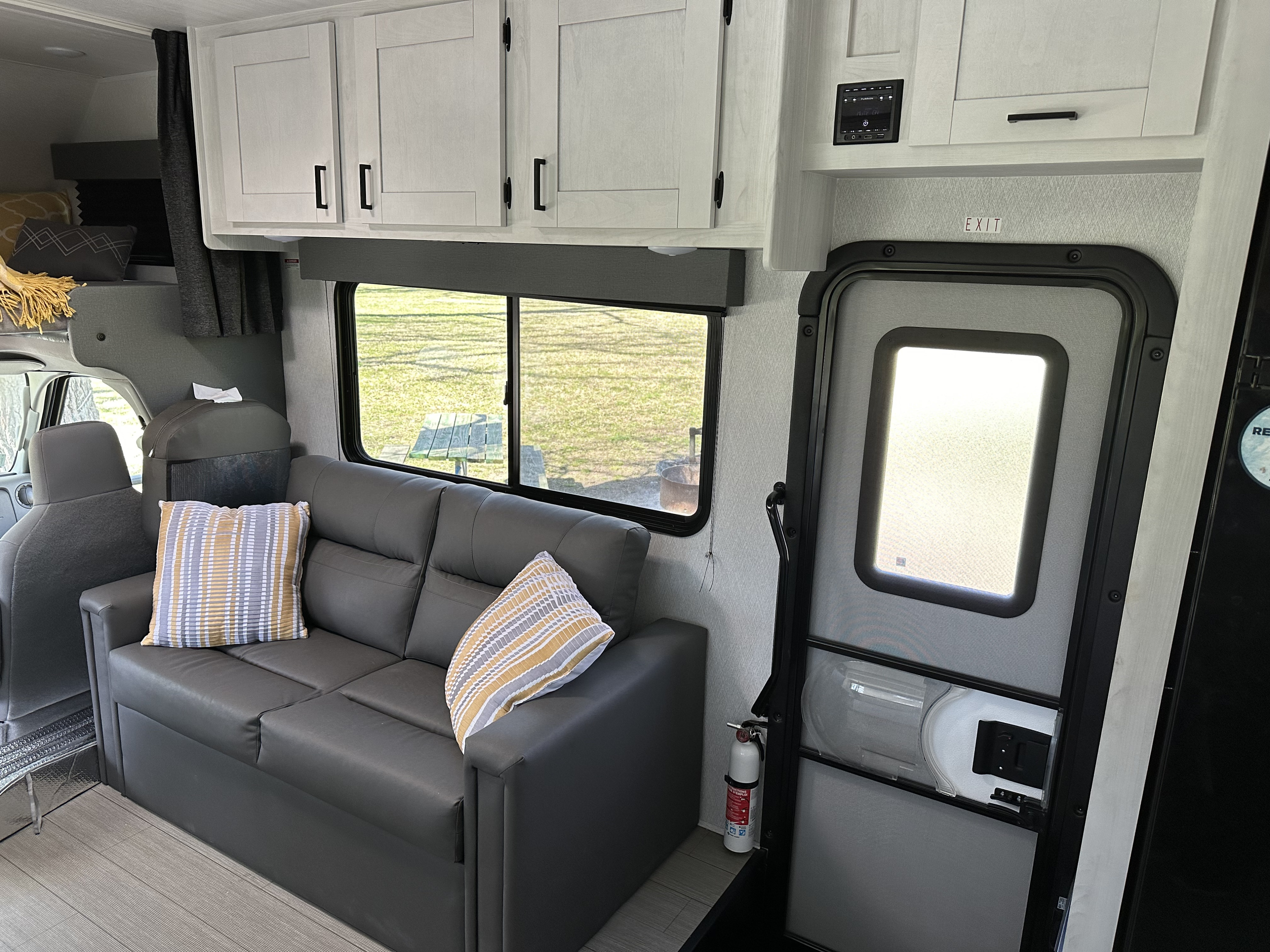 An RV rental interior with a gray sofa, light cabinets, a window looking outside, and an exit door. A fire extinguisher is by the door.. Forest River 3100FB 2024
