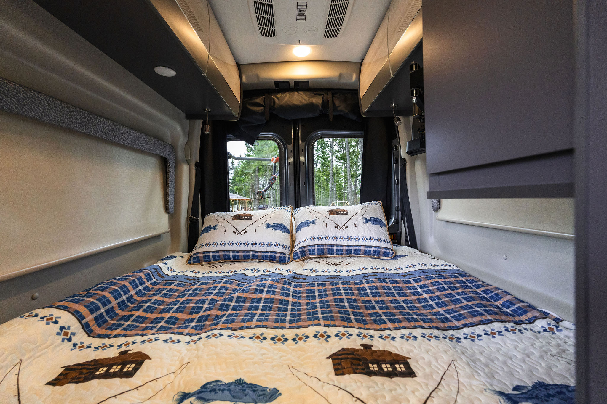 Experience comfort in this RV rental bedroom with fishing-themed quilt & pillows. Enjoy forest views from your camper van.. Thor Sanctuary 2023
