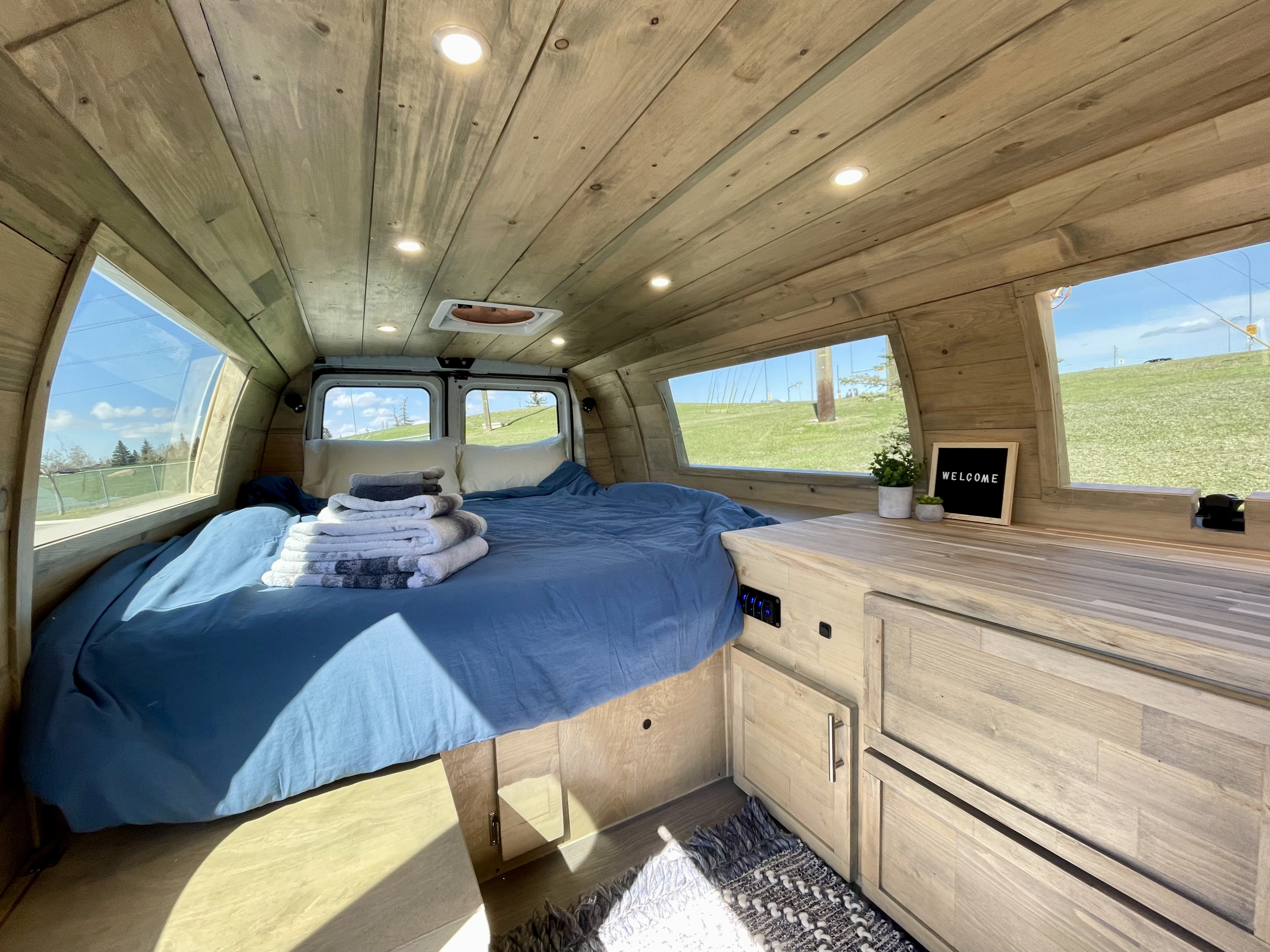 Cozy RV rental interior with light wood paneling, a comfy blue bed, and large scenic windows. Perfect for your trip.. Ford Ford E350 2011