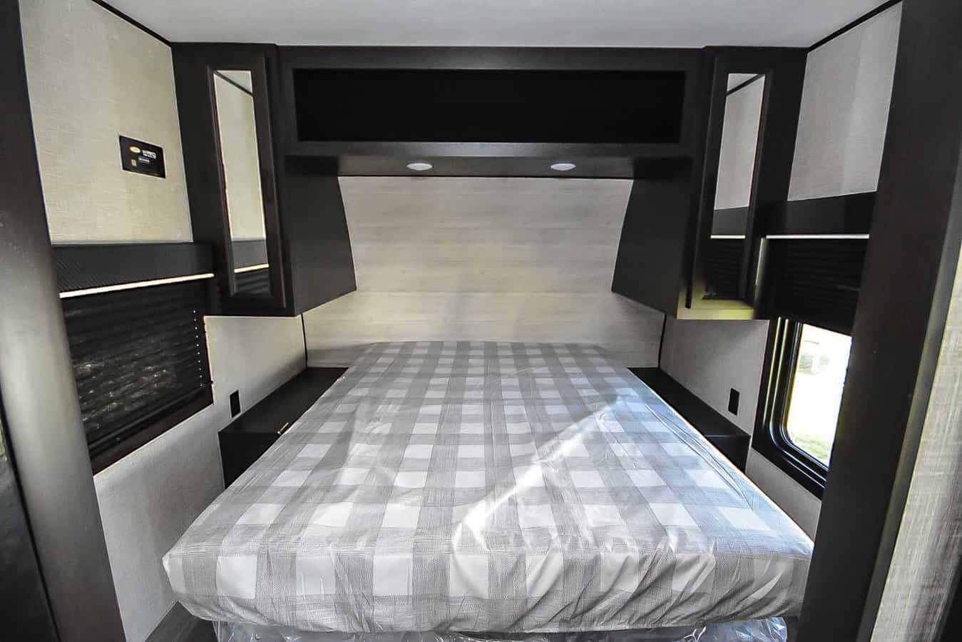 Clean RV rental bedroom with a gray plaid mattress, dark overhead cabinets, side tables, and window blinds.. Jayco Jay Flight 2023