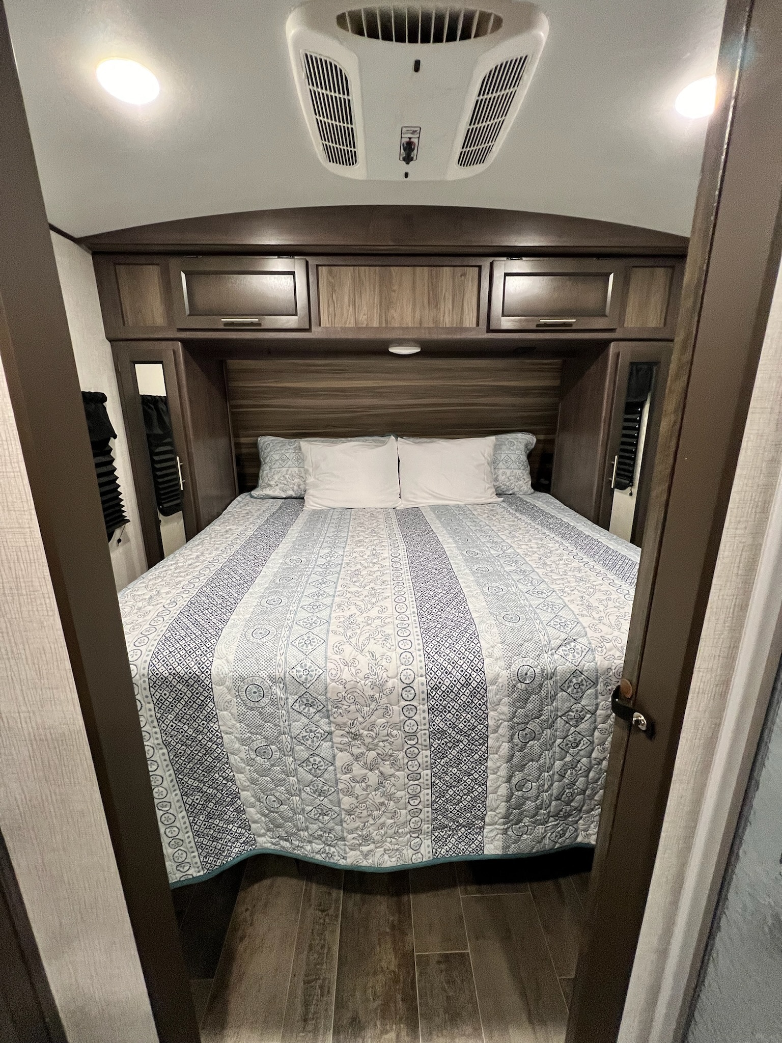 This RV rental bedroom features a comfy bed with a blue/white quilt, wood cabinets, and an overhead AC unit.. Crossroads Sunset Trail 2021