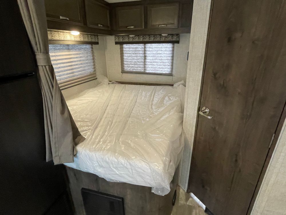 Inside an RV rental, showing a bed with a plastic-covered mattress, overhead cabinets, and windows with blinds.. Gulf Stream Conquest 2023