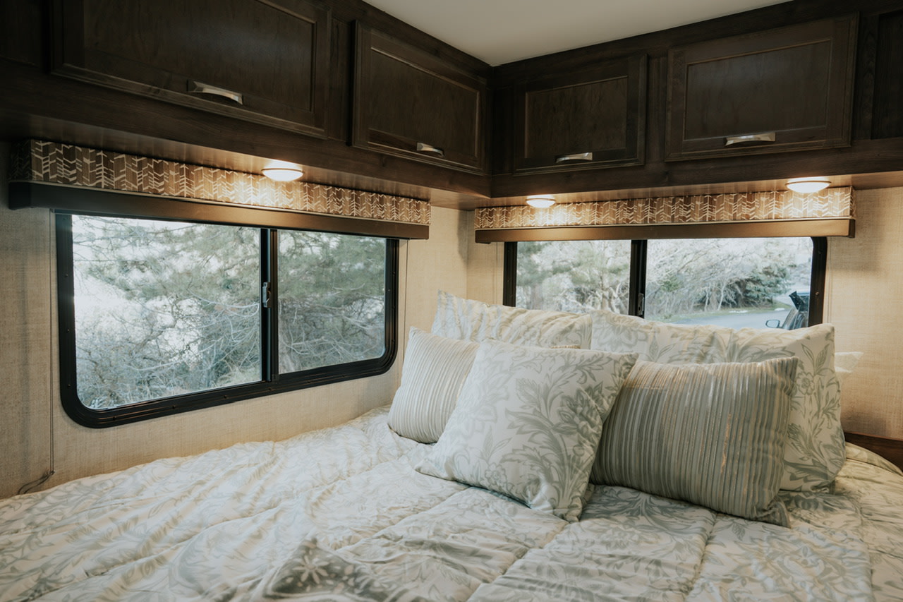 Relax in this cozy RV rental bedroom. Features a comfortable bed, overhead cabinets, and scenic window views.. Gulf Stream Conquest 2023