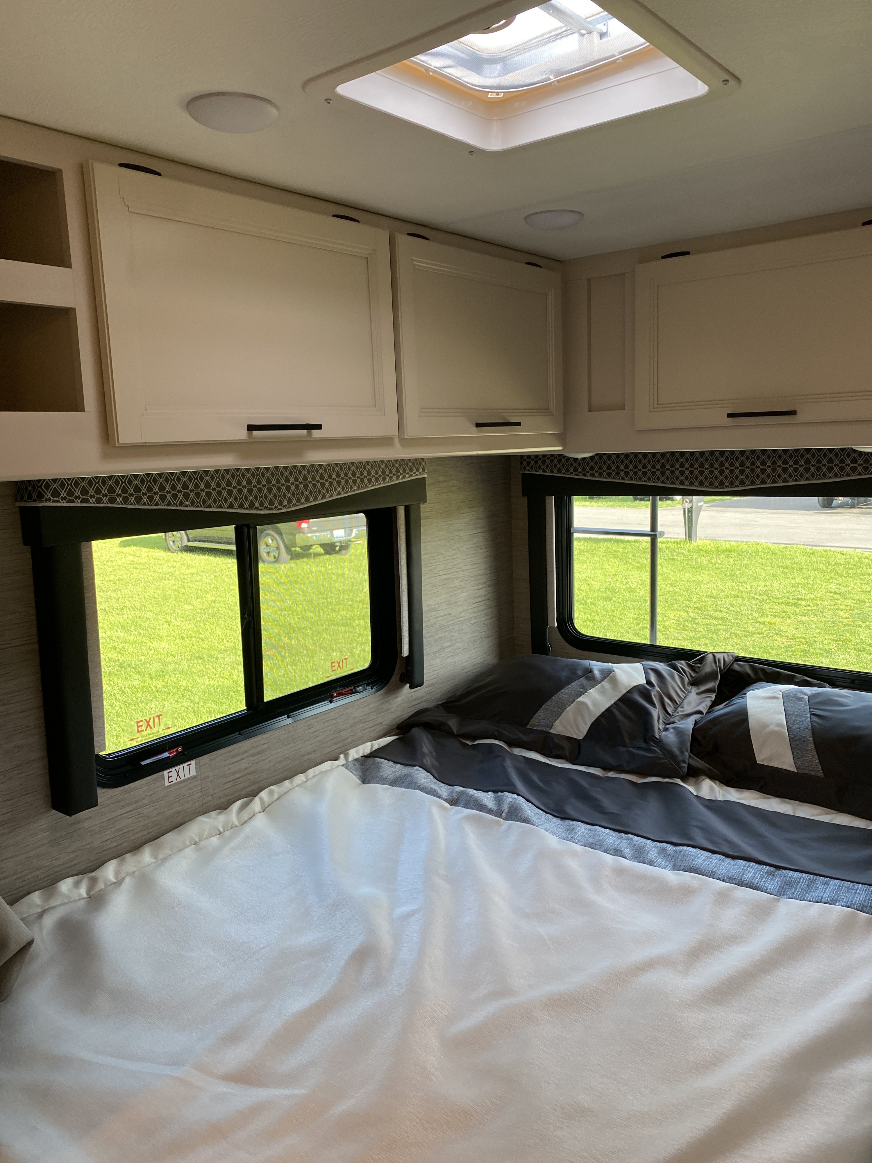Clean RV rental bedroom with comfortable bedding, overhead cabinets, a skylight, and large windows looking out to nature.. Ford F350 22E MC 2023