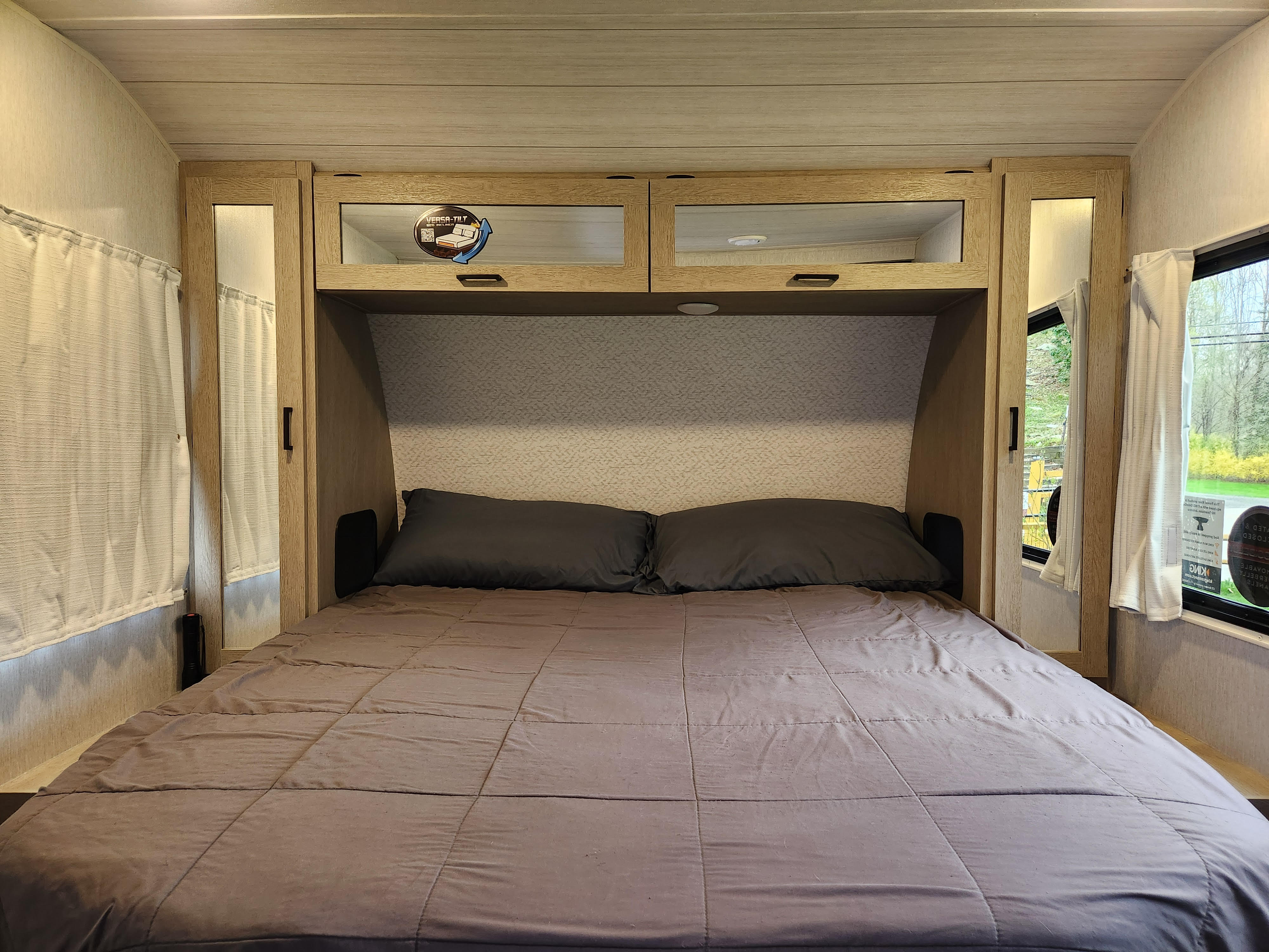 Cozy RV rental bedroom features a gray bed, overhead cabinets with a 'VERSA-TILT' sticker, and side windows with curtains.. Forest River Wildwood 2024