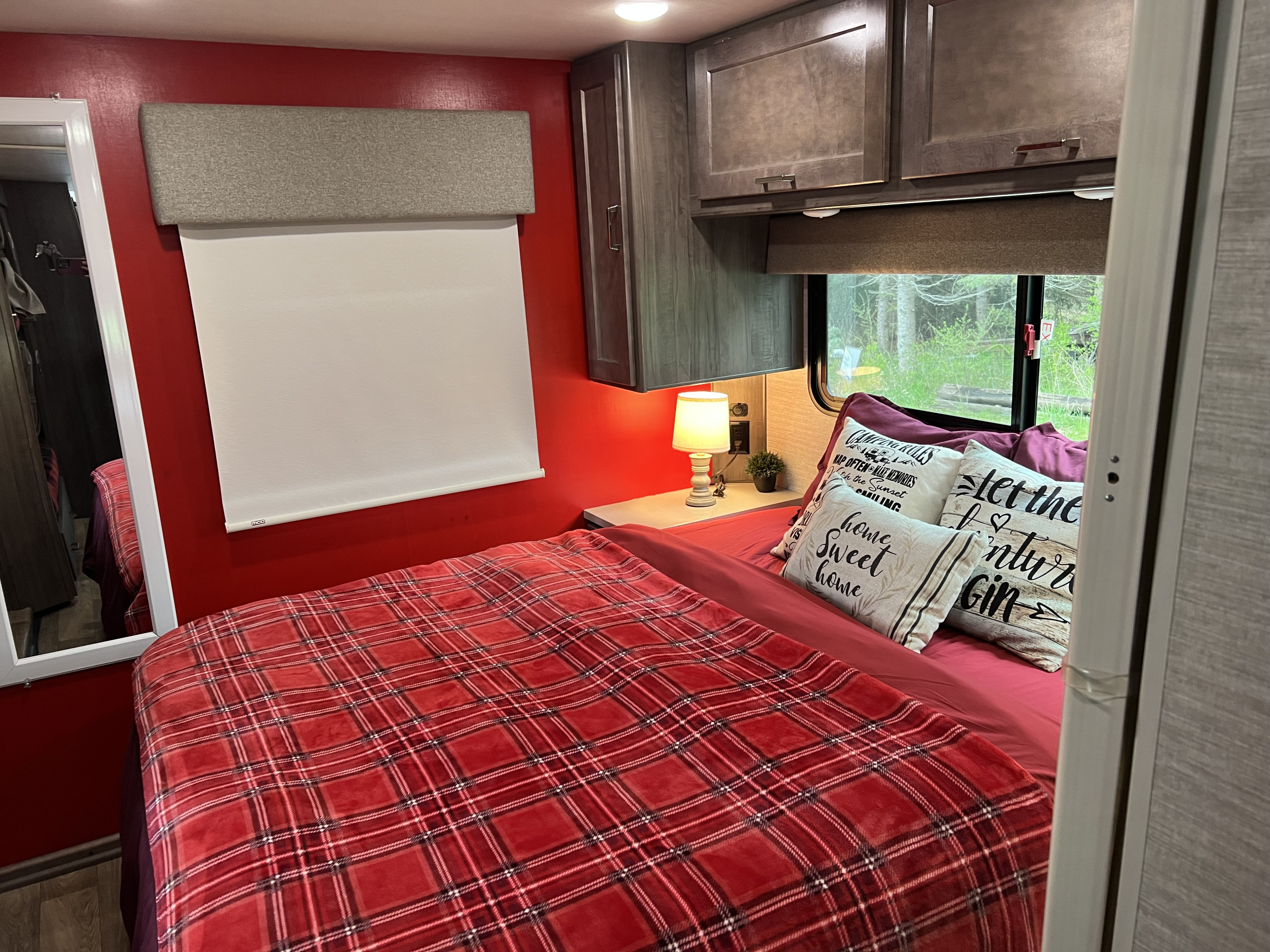 Inside a vibrant RV rental: red bedroom with plaid blanket, cozy pillows, bedside lamp, and overhead cabinets.. Winnebago Minnie Winnie 2020