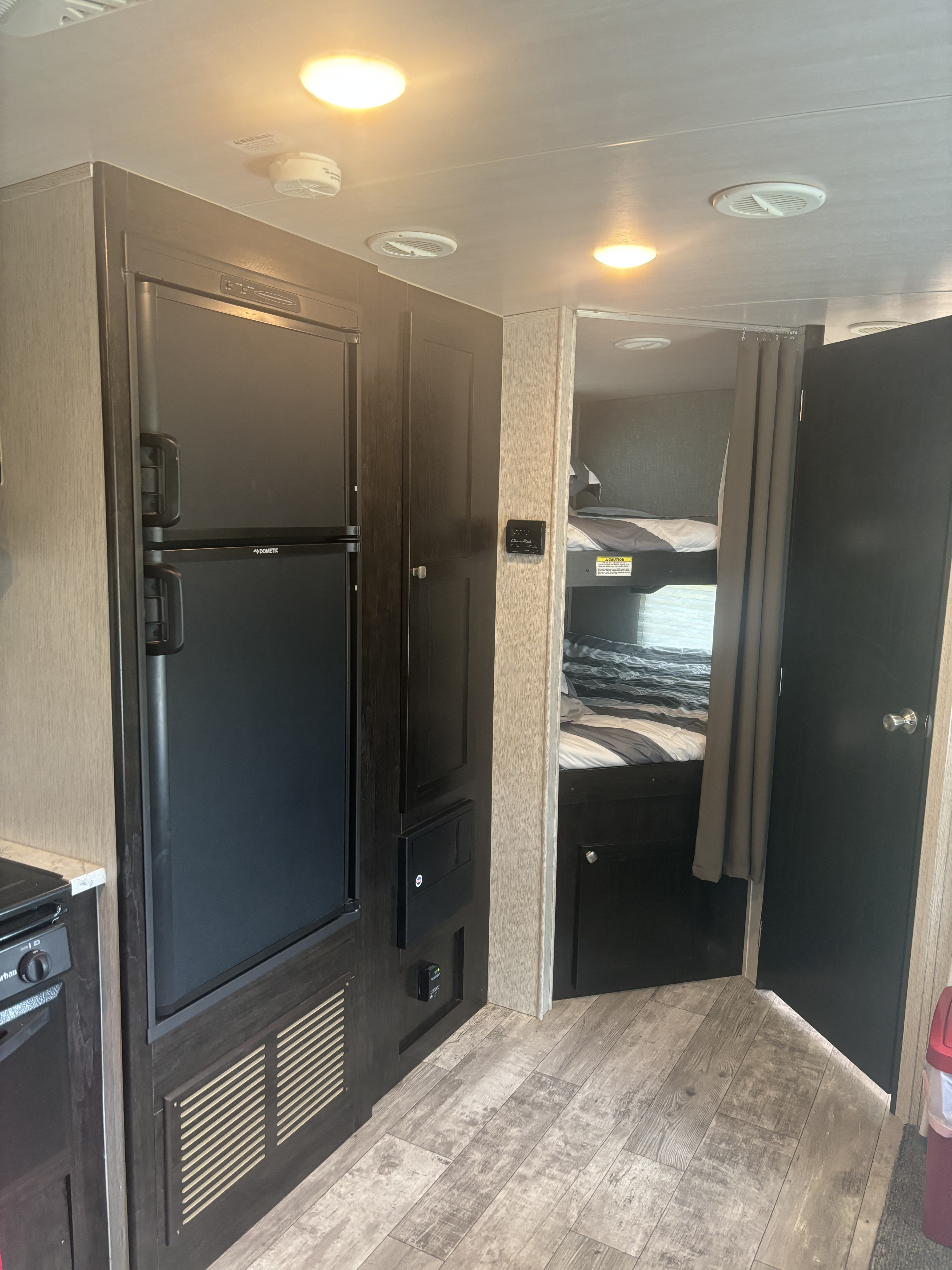 RV rental interior: full-size fridge, storage, and entrance to bunk beds with privacy curtain. Features modern lighting, vents, and wood-look flooring.. Heartland RVs North Trail 2018