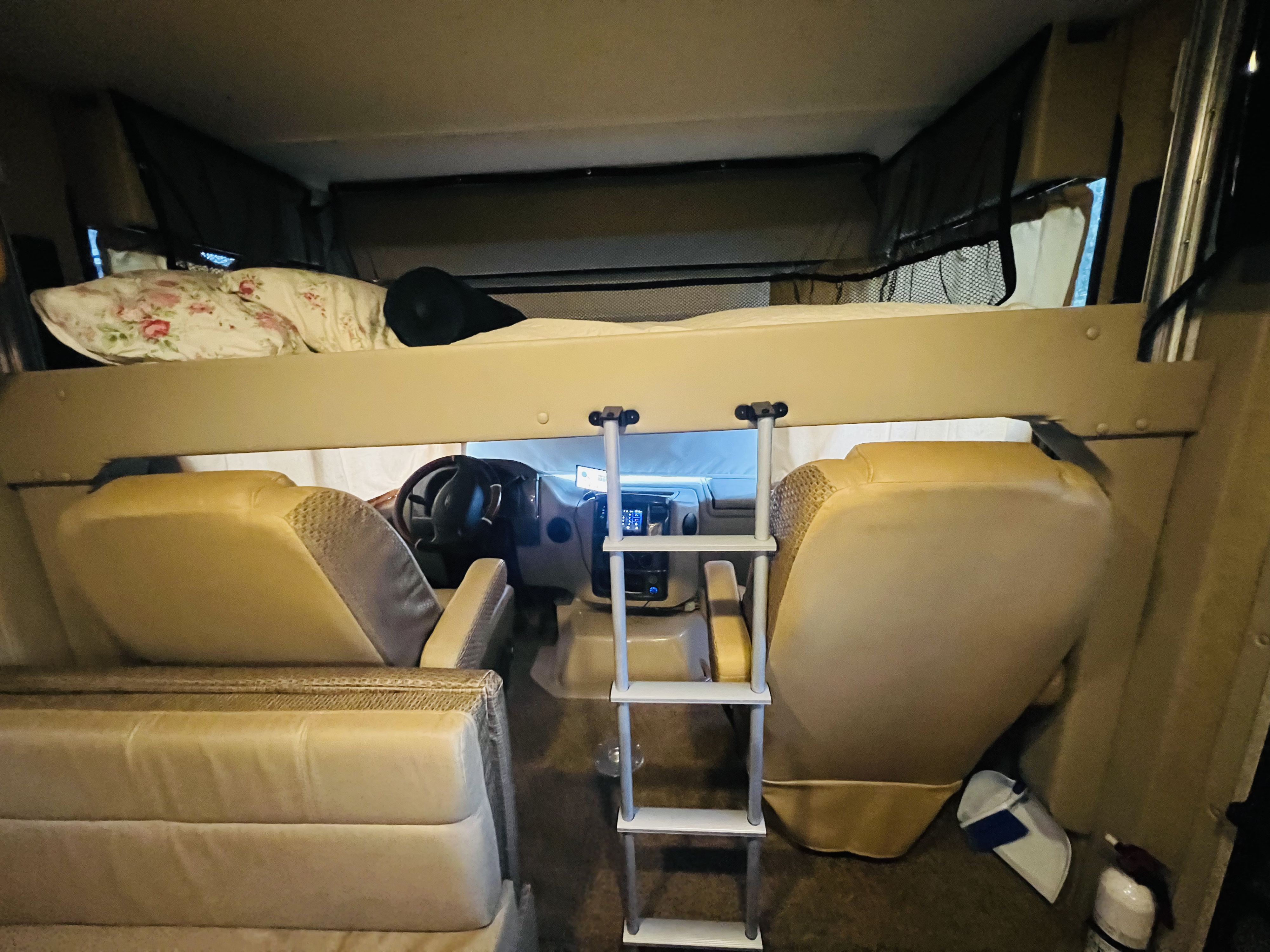 Inside an RV rental: a comfortable over-cab bunk with a ladder, driver's cockpit, and lounge seating for travel.. Thor Vegas 2016