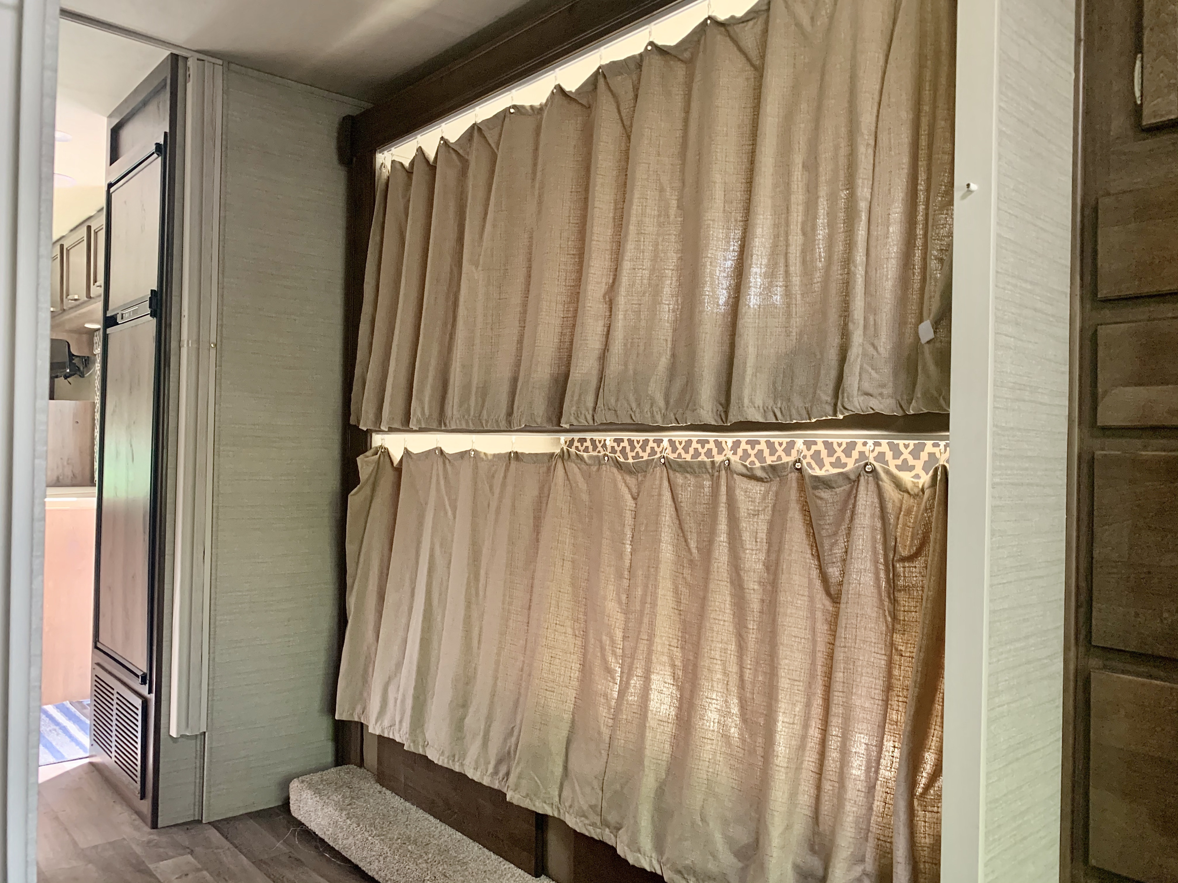 Inside an RV rental: two levels of light privacy curtains for bunk beds, with a refrigerator and cabinets visible on the sides.. Jayco Redhawk 2019