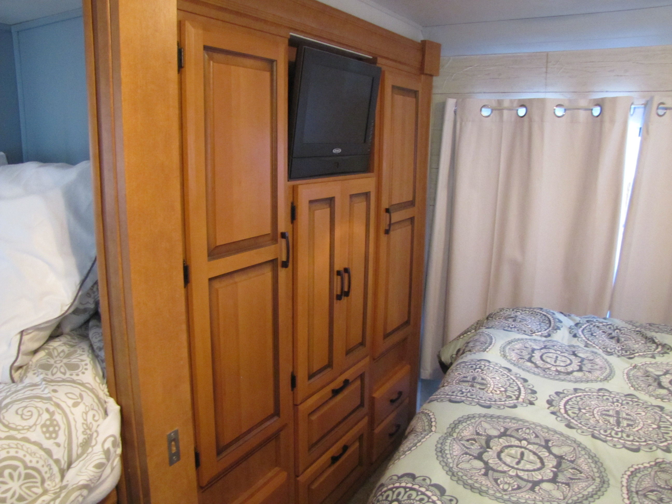 This RV rental bedroom features light wood cabinetry, an integrated TV, and a comfy bed with patterned comforter and curtains.. Forest River Sunseeker 2011