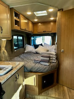 Explore this RV rental's cozy interior, featuring a made bed in the bedroom, wooden kitchen cabinets, and a bathroom door.. Thor Four Winds 2021