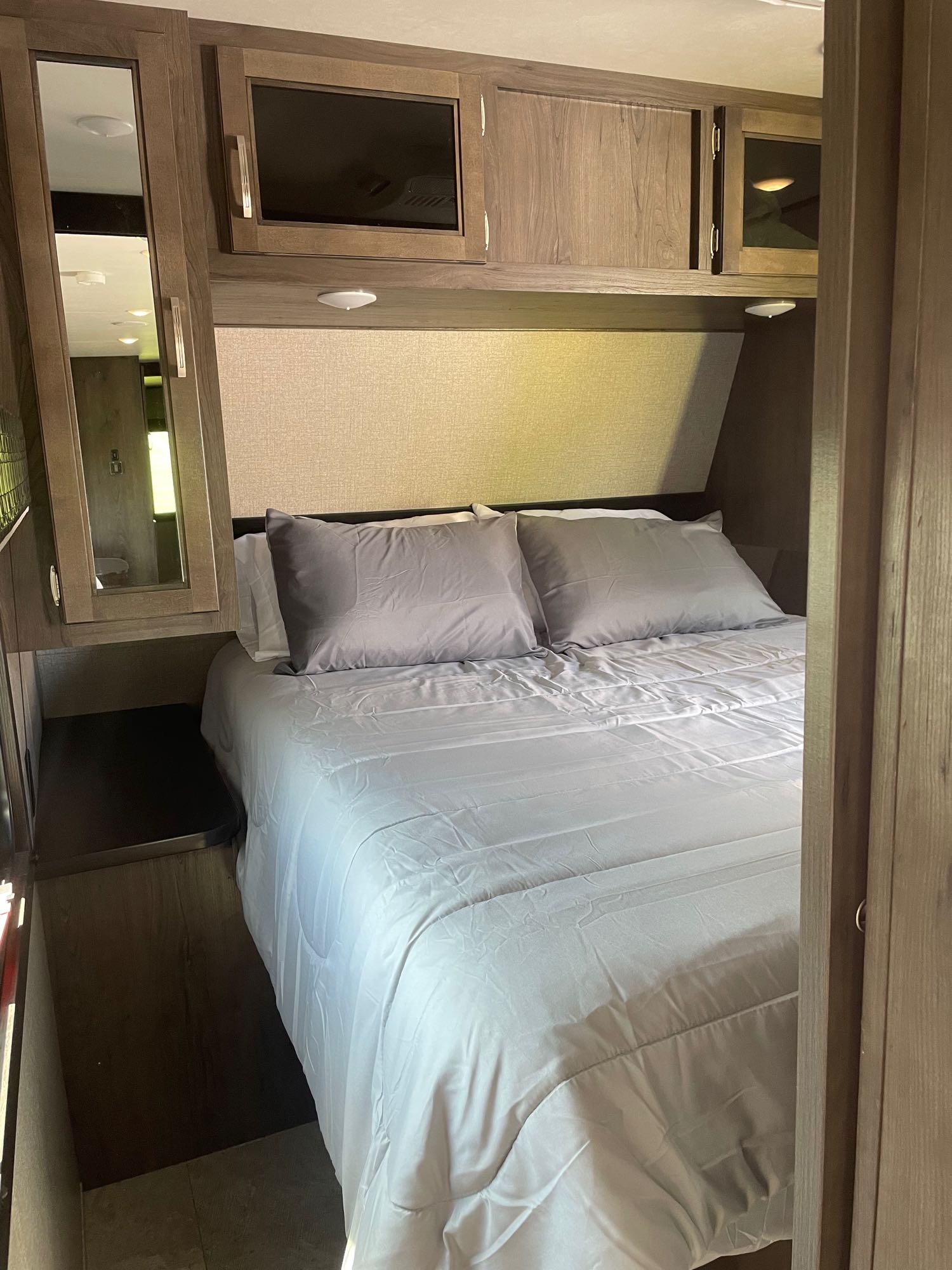 RV rental bedroom view: comfortable bed with gray bedding, overhead wood cabinetry including a TV, mirror, and nightstand.. Jayco Jay Flight 2020