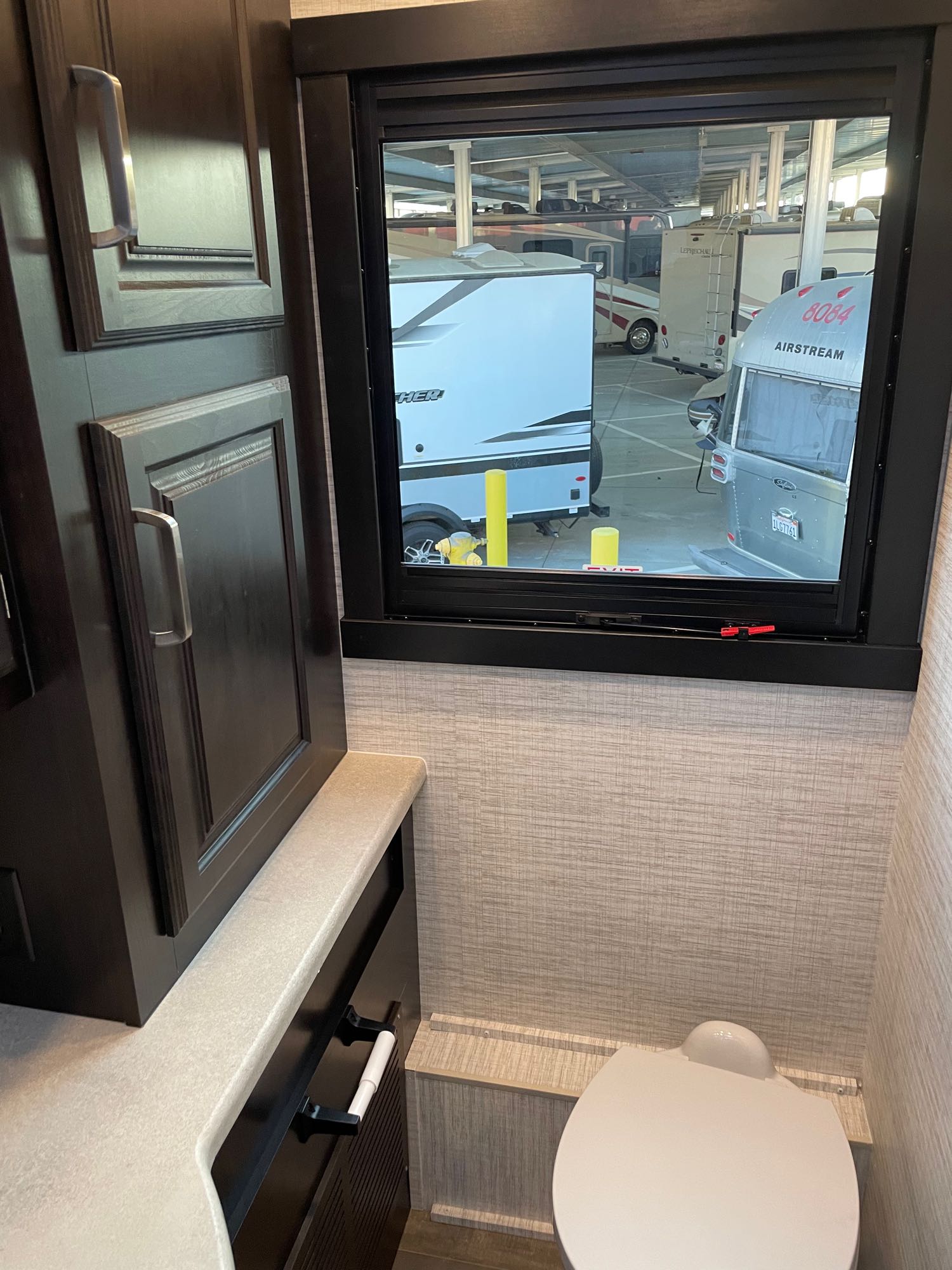 Inside an RV rental: a compact bathroom with toilet and window view of other parked RVs. Dark wood cabinets and light walls.. Ford Entegra Vision XL 36C 2024