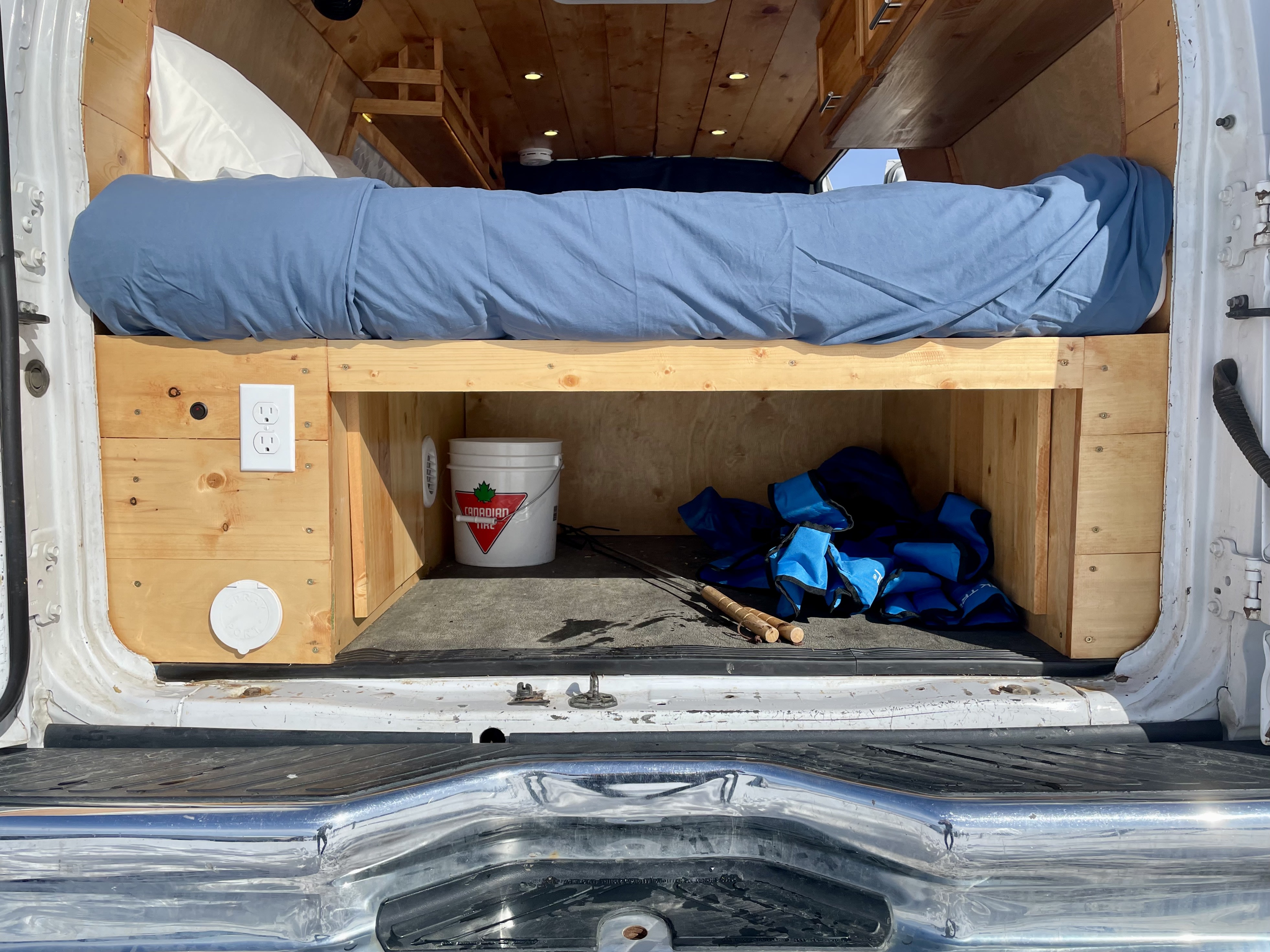 Inside a DIY RV rental: comfy bed, under-bed storage with bucket, blue gear, & skewers. Power outlets & wood ceiling.. Gmc Savanna 2017