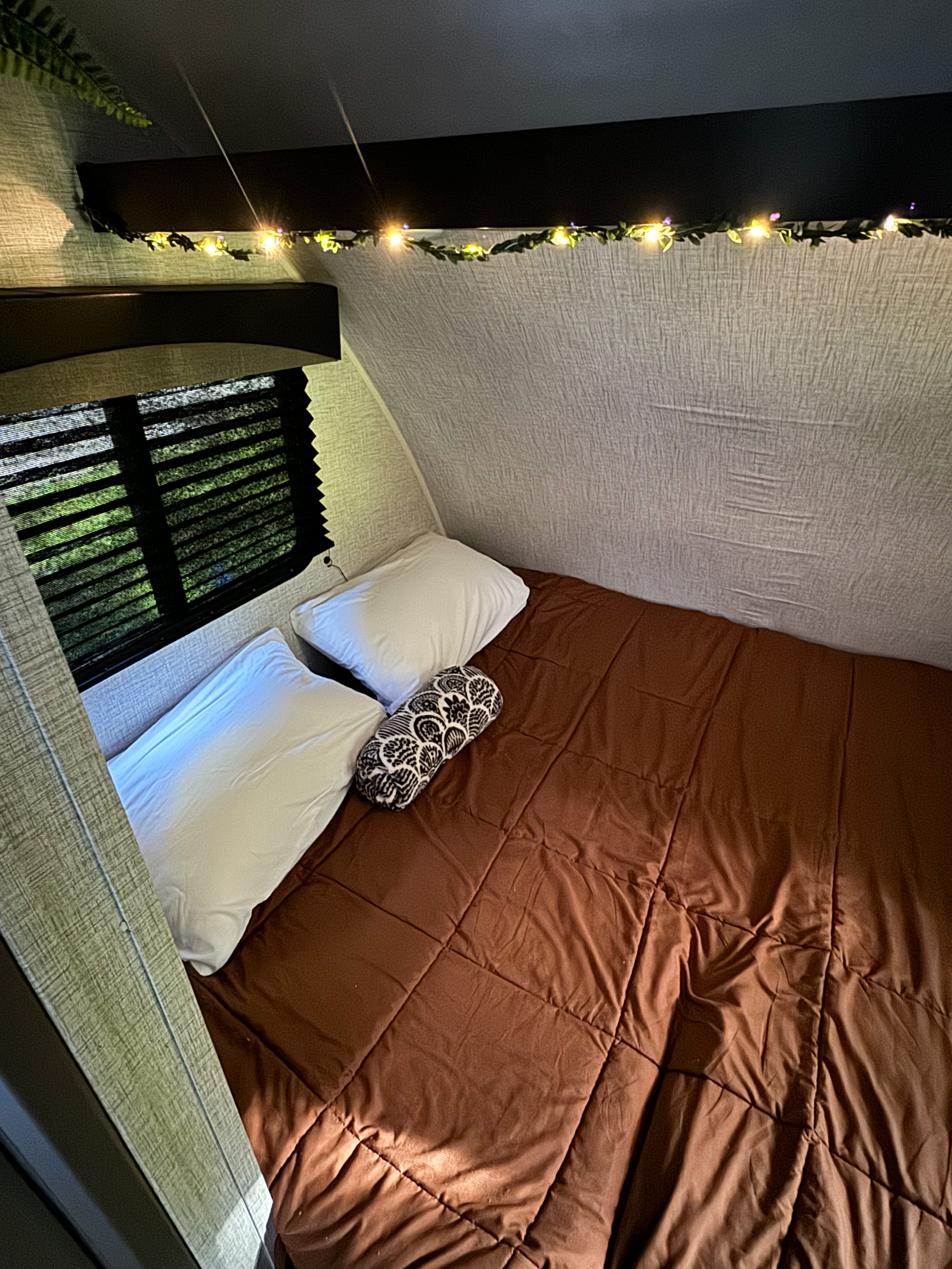 Cozy RV rental bedroom with brown comforter, white pillows, and fairy lights. Window shows greenery.. Braxton Creek Bushwacker Plus 17BH 2023