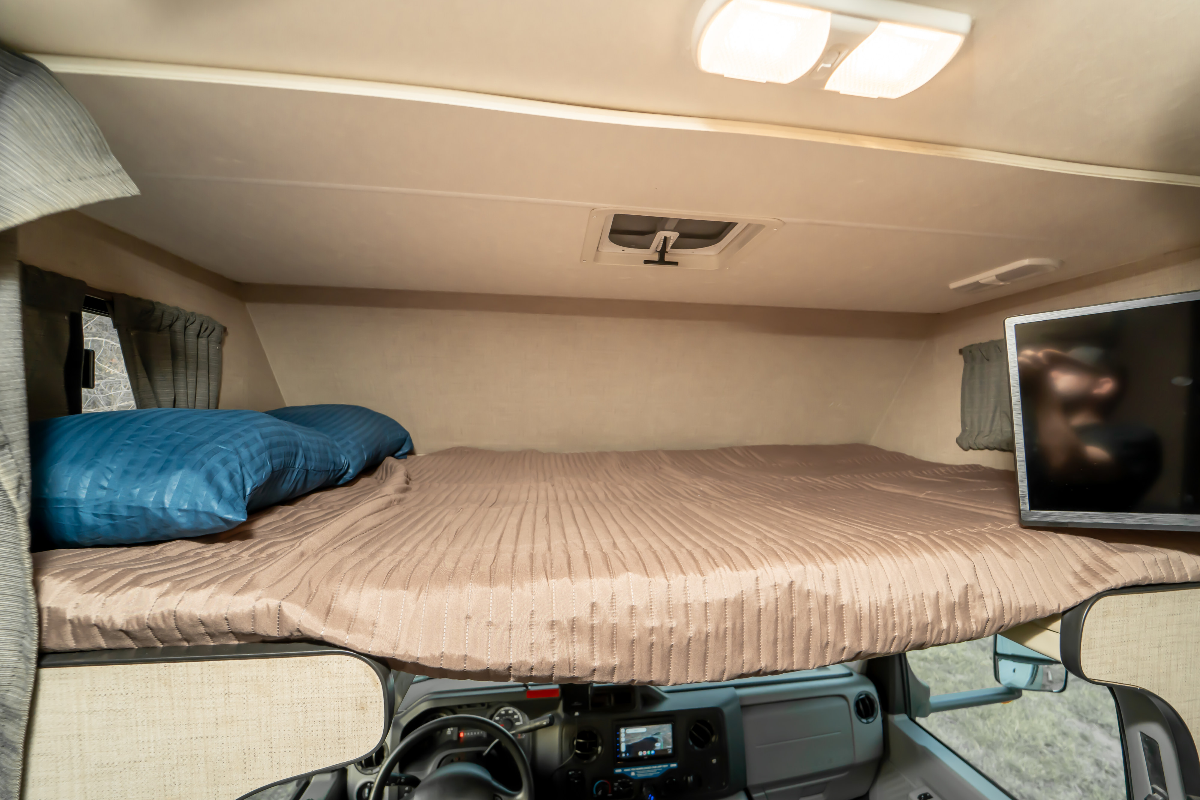 Interior of an RV rental showing the cab-over bed, fitted with a brown quilted comforter, blue pillows, and a TV.. Thor Majestic 2019