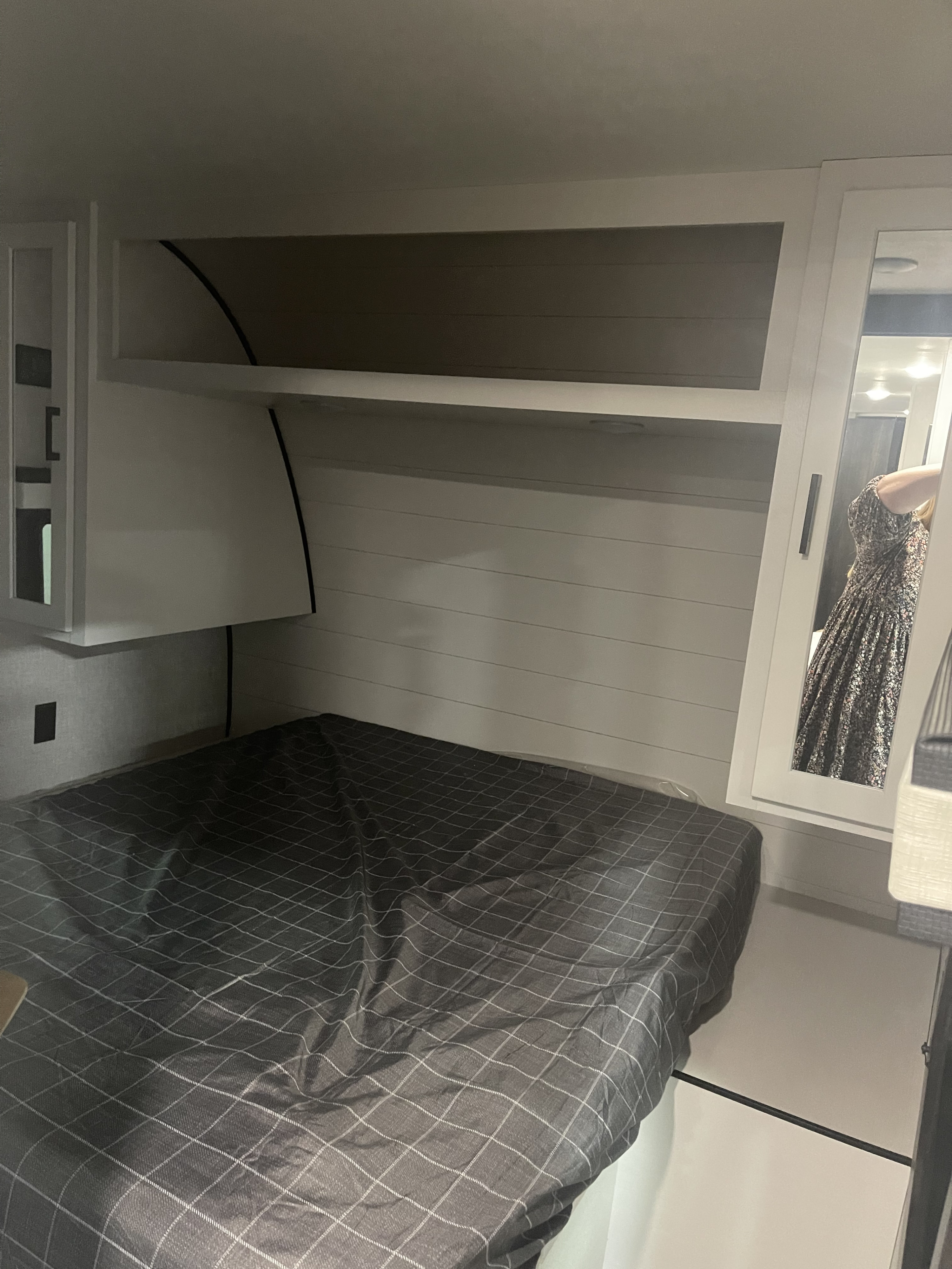 Inside an RV rental: a cozy bedroom with a dark grey patterned bed, overhead cabinets, and a convenient mirror.. Jayco JayFlight 2024