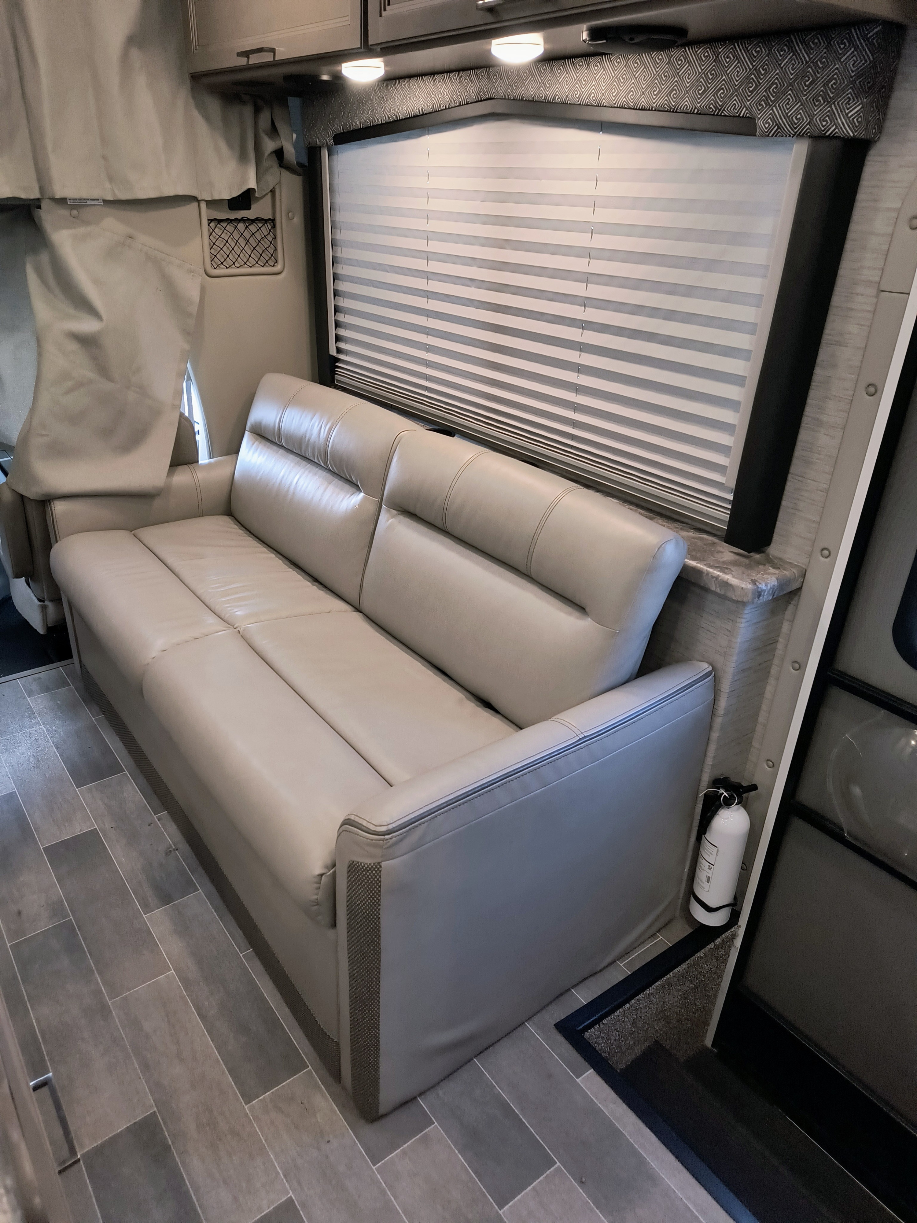 Inside an RV rental: a modern living area featuring a light leather sofa, window blinds, and tiled floor. Fire extinguisher visible.. Thor Motor Coach Mgnitude 2022