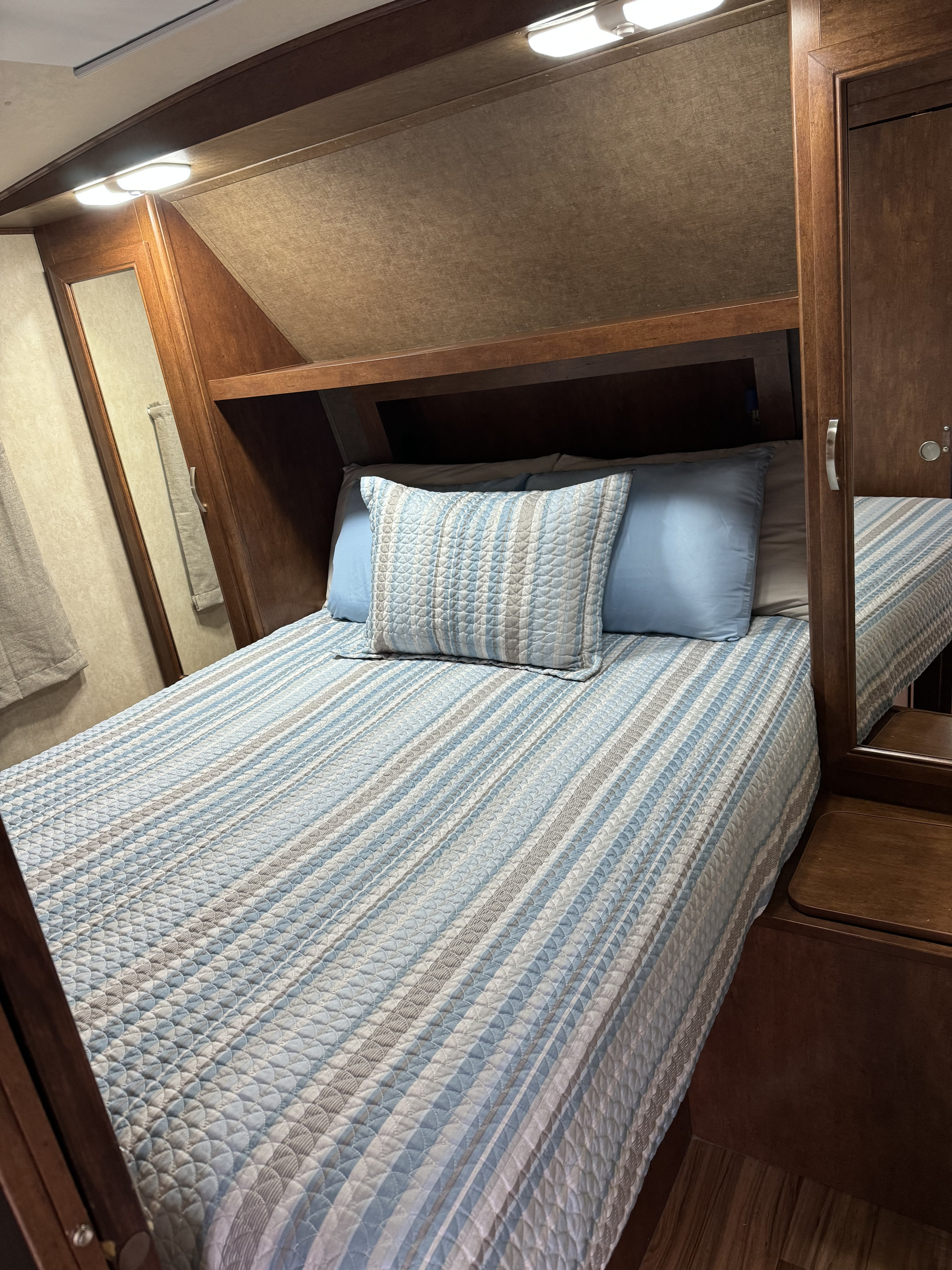 Cozy RV rental bedroom features a bed with striped blue and grey bedding, bright overhead lights, and wooden built-in storage.. Forest River Vibe Extreme Lite 2015