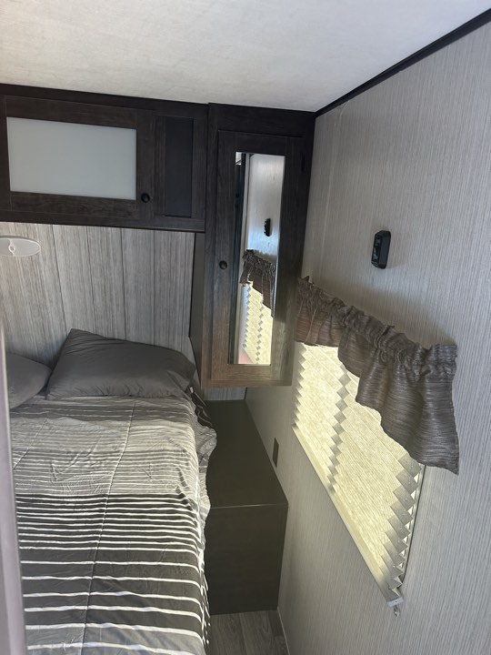 Inside a functional RV rental bedroom: bed with grey stripes, built-in storage, and window with blind.. Heartland RVs Pioneer 2020