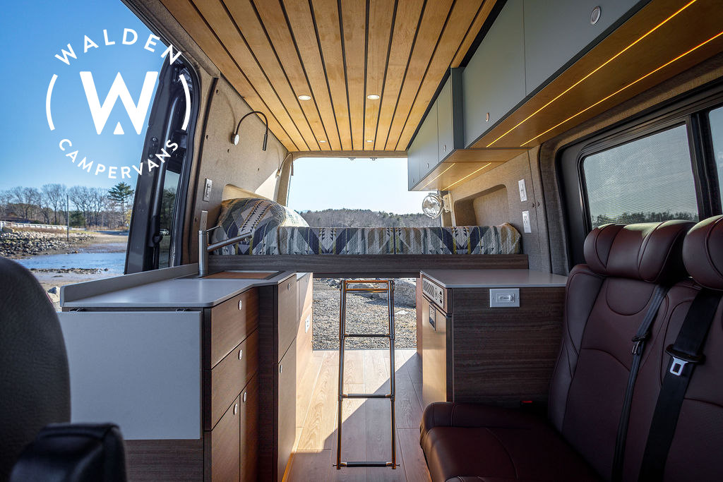 Modern RV rental interior of a custom camper van. Features include a bed, kitchen, wood ceiling, and scenic open rear view.. Mercedes-Benz Sprinter 2023