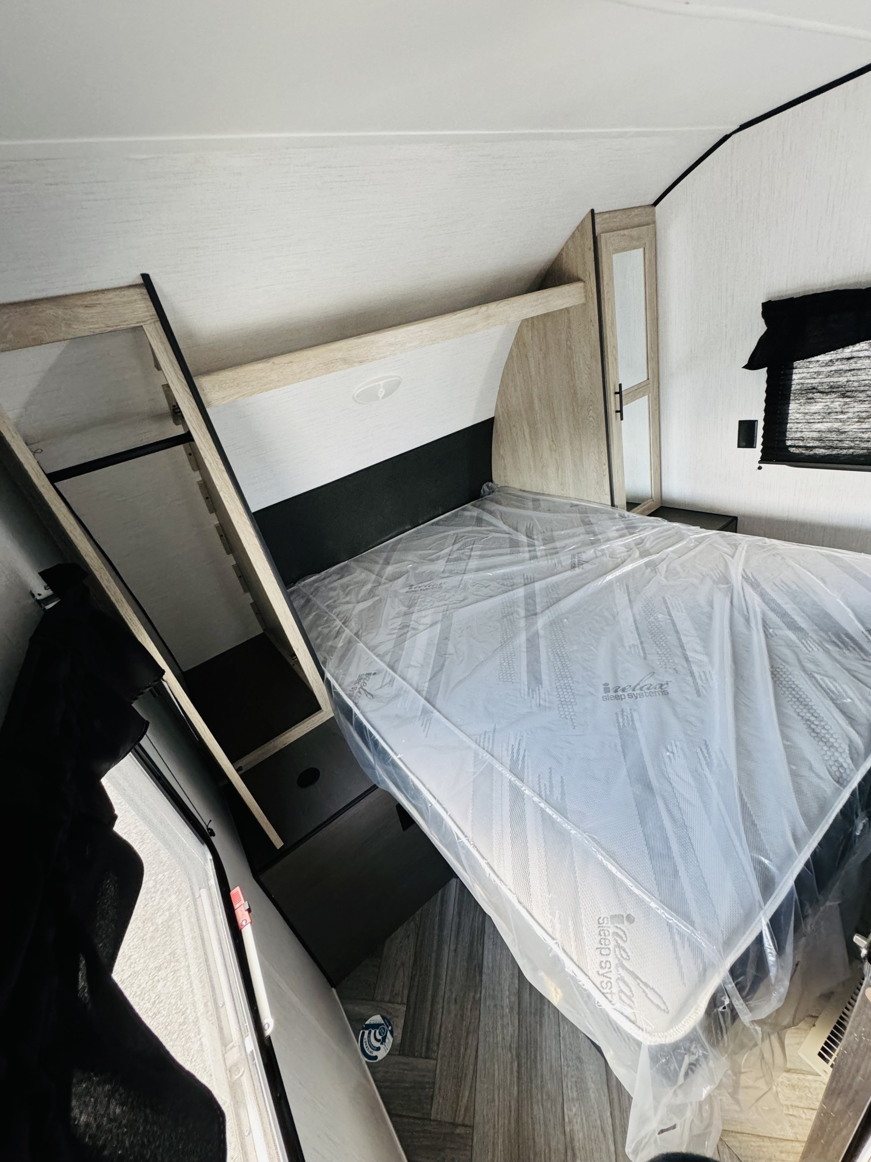 This RV rental bedroom features a new, plastic-wrapped i-relax mattress, shelving, and a window.. Heartland RVs Prowler 2024