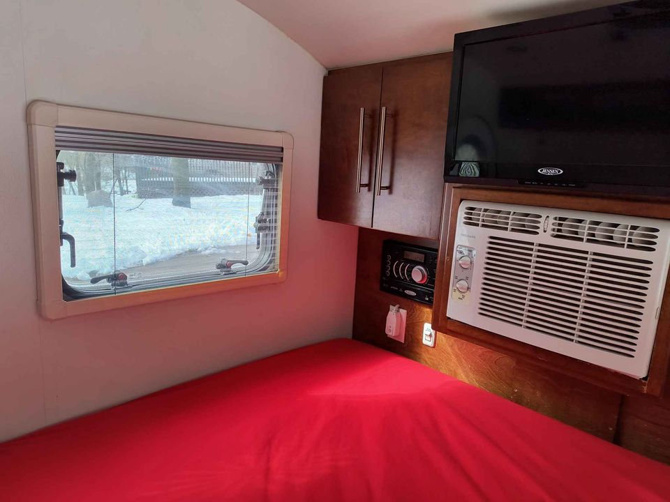 Cozy RV rental bedroom with a vibrant red bed, window viewing snow, TV, AC unit, and wooden storage cabinets.. T@G T@G Trailer 2015