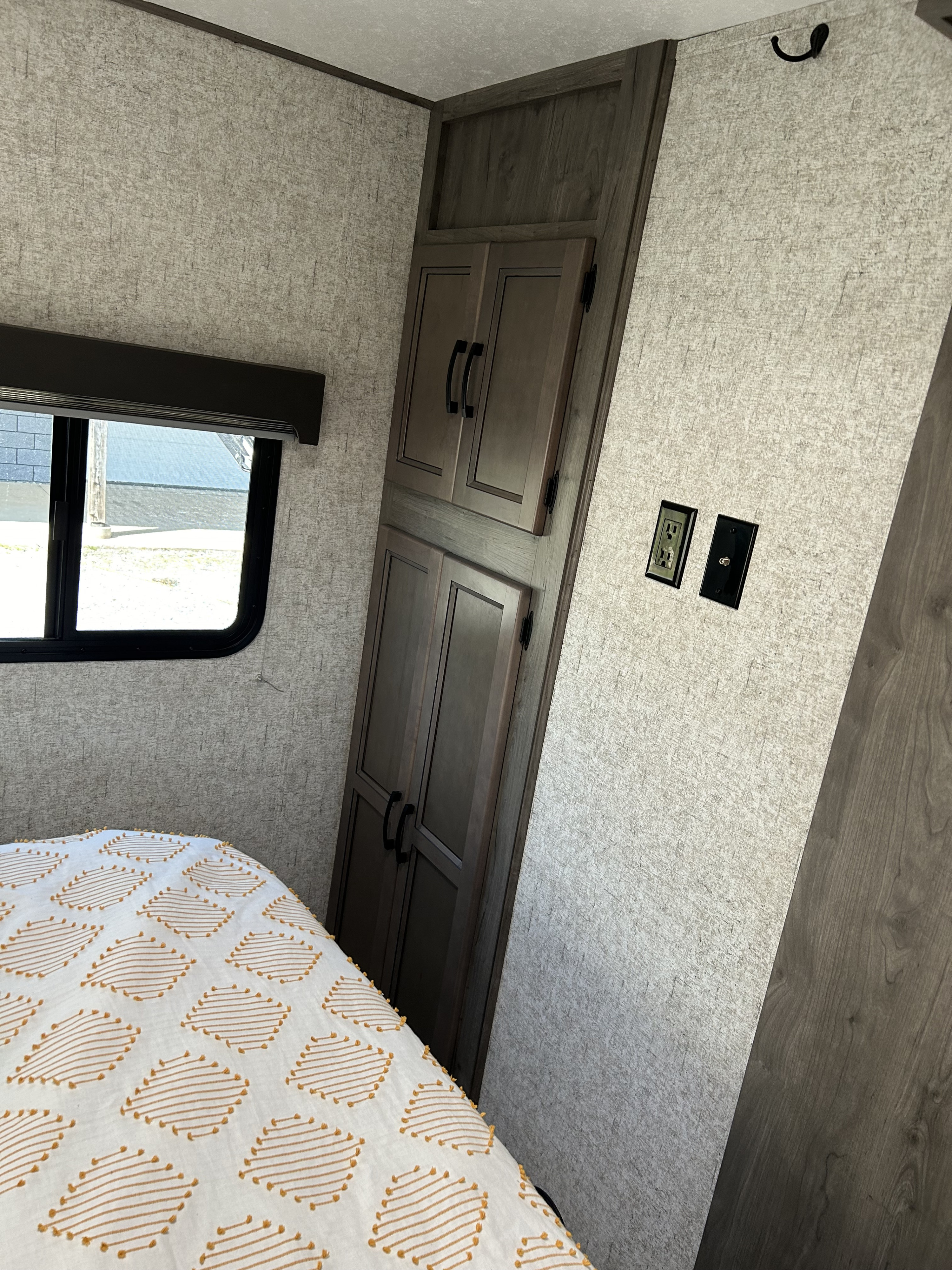 RV rental interior: bedroom features patterned bed, window, and dark wood storage. Textured walls, outlets, & a hook. Comfortable travel.. Coachmen Apex 2021