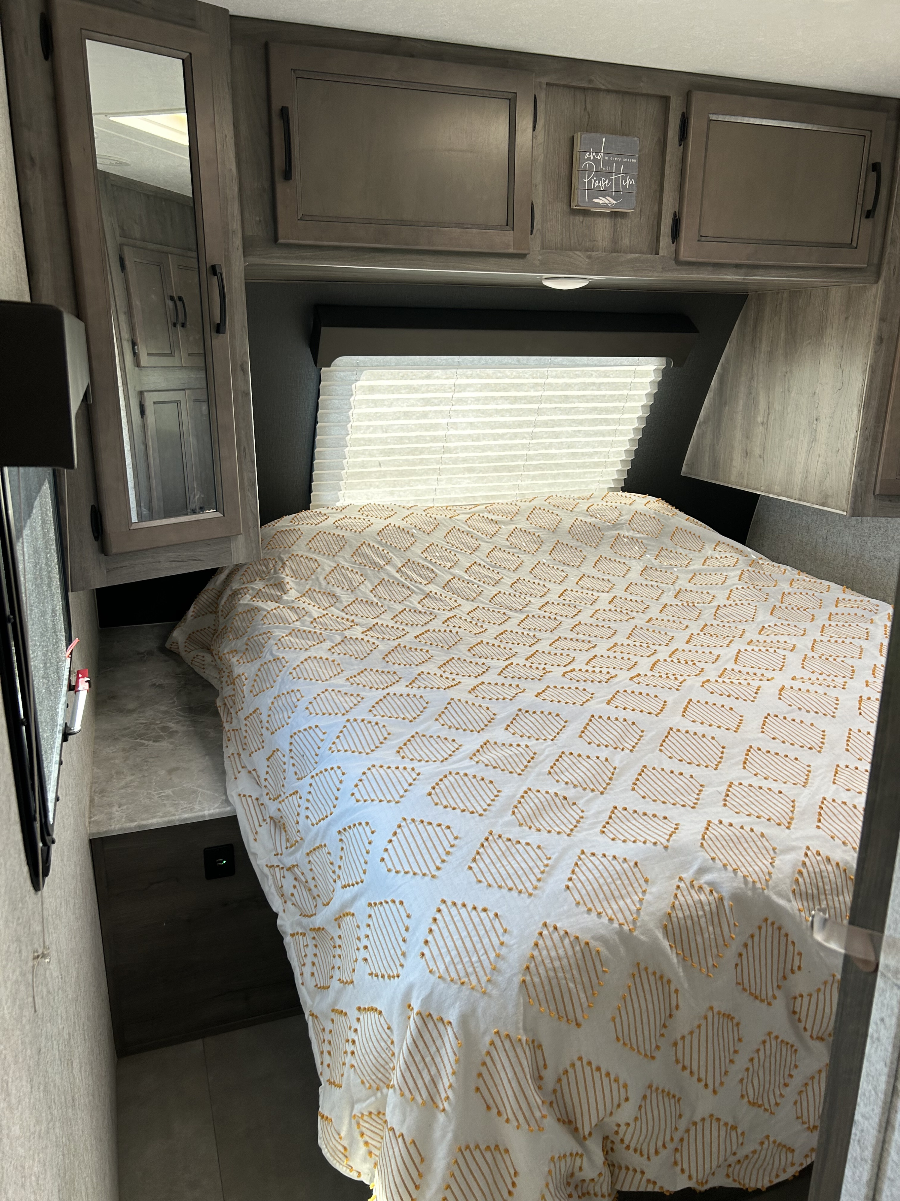 Inside an RV rental, a compact bedroom features a bed with a patterned comforter, overhead cabinets, and window blinds.. Coachmen Apex 2021